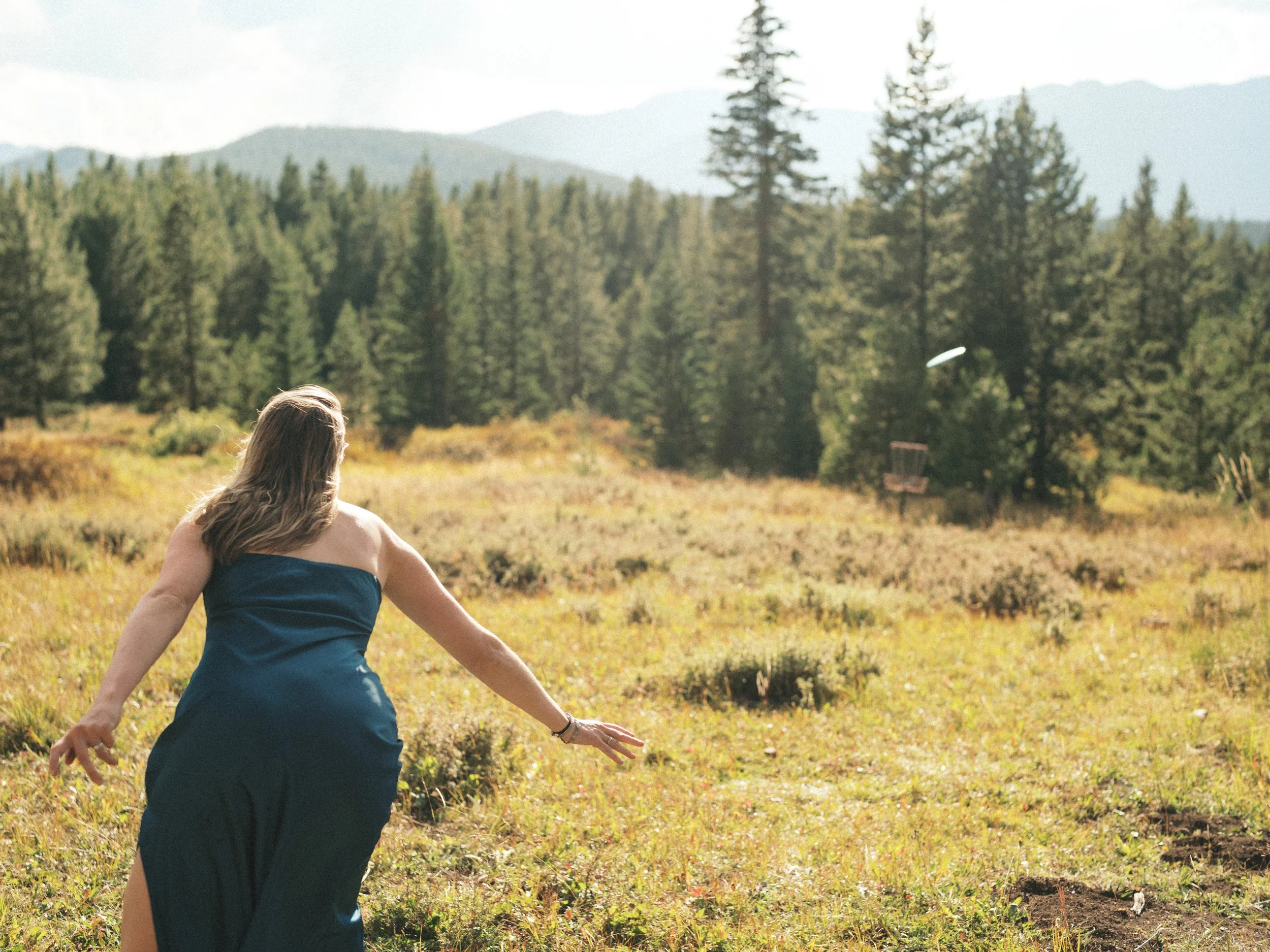 A woman with long hair wearing a blue strapless dress, sitting in a field of grass and wildflowers with a backdrop of pine trees and distant mountains.
