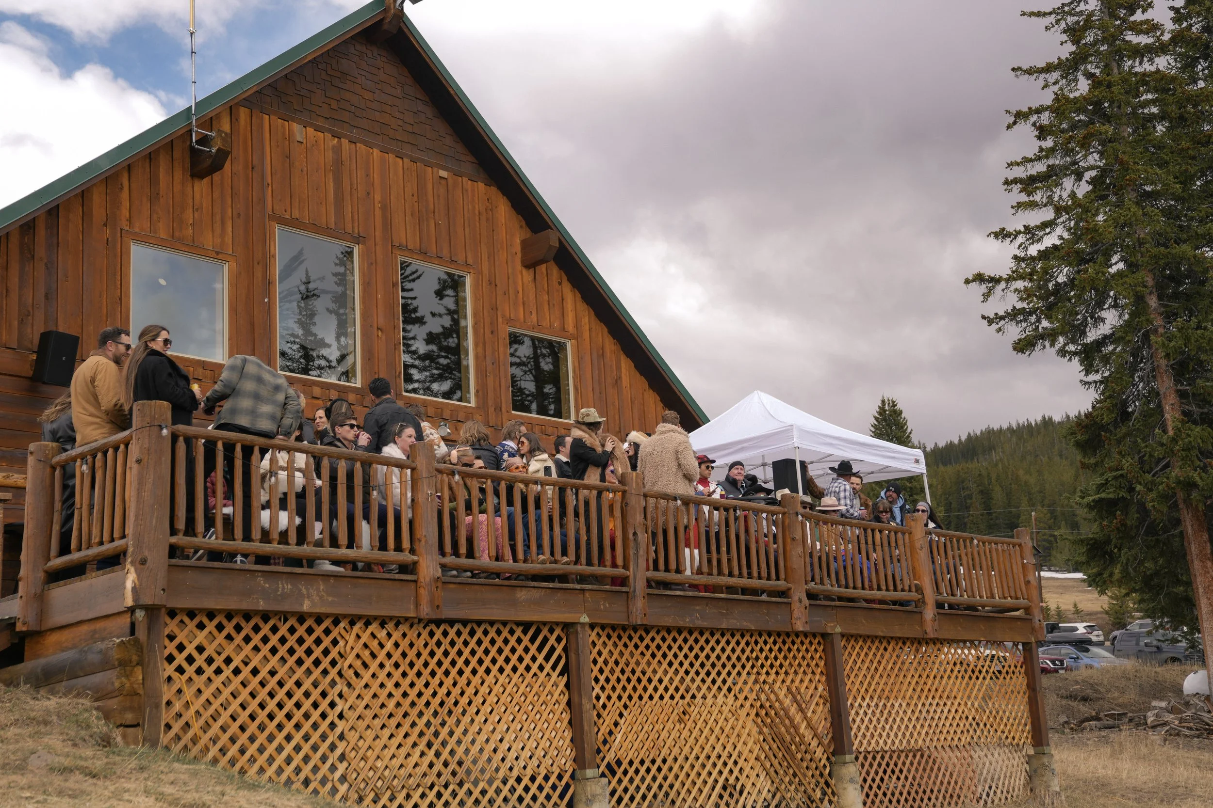 People gathered on a wooden deck outside a large wooden cabin, with trees and cloudy sky in the background.