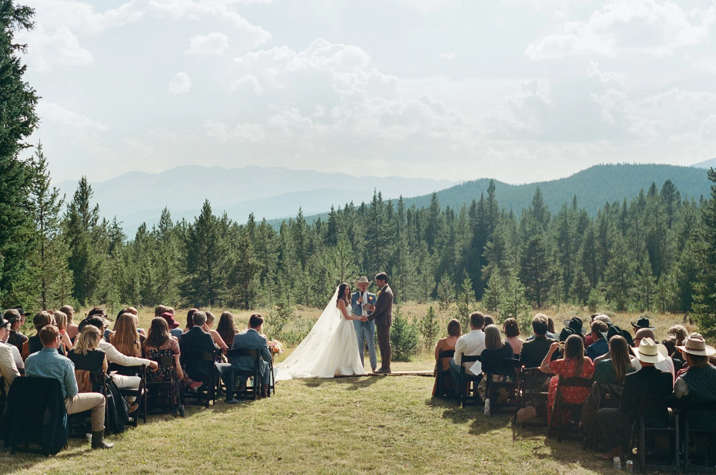 An outdoor wedding ceremony in a forested mountainous area with guests seated watching a bride and groom exchange vows, officiated by a person holding a microphone. The bride wears a white gown with a long train, and the groom wears a dark suit. A large crowd sits on chairs among lush green trees with mountains and a partly cloudy sky in the background.