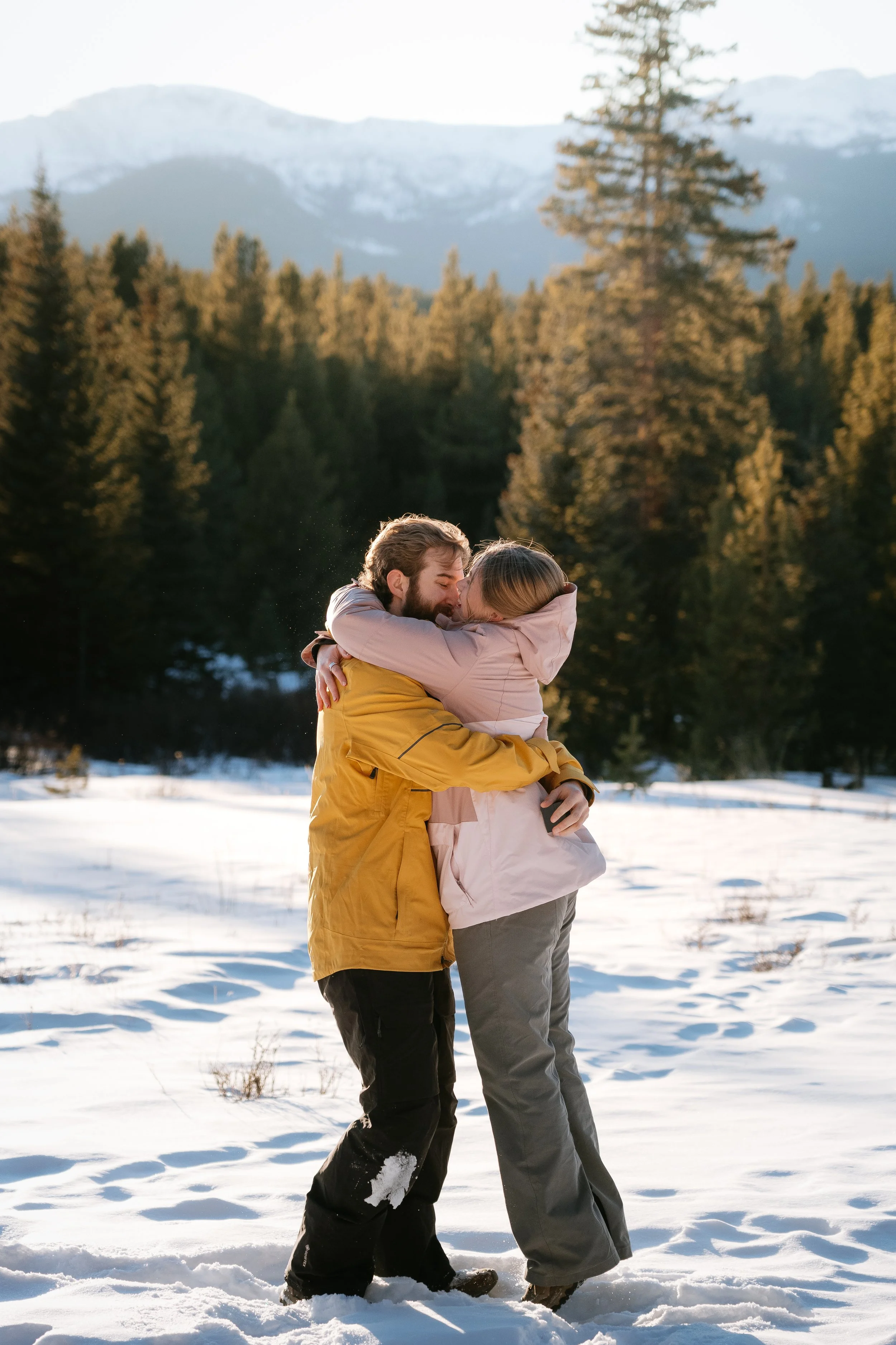 A couple hugging in a snowy outdoor setting with trees and mountains in the background.