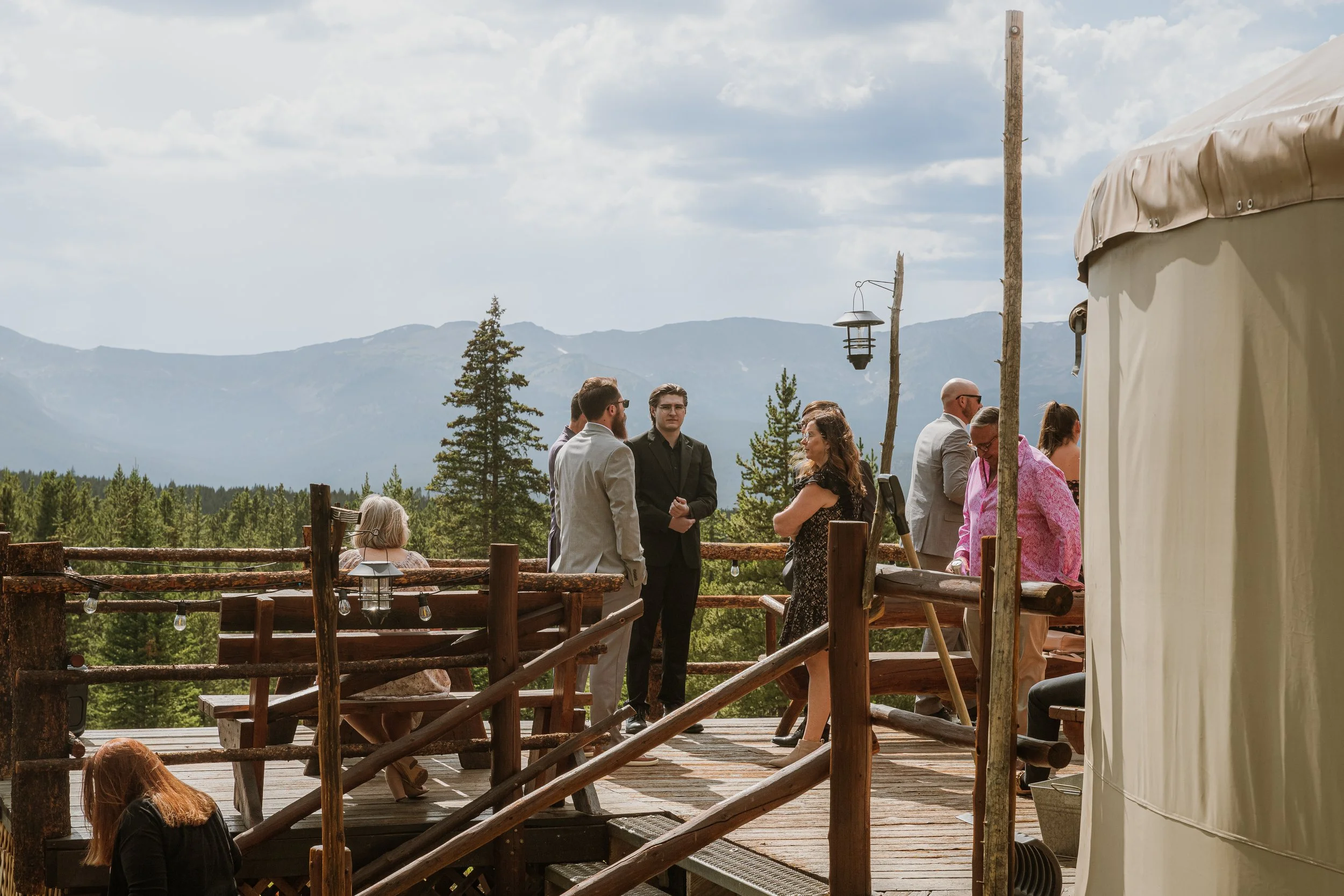 People gathered outdoors on a wooden deck with mountain scenery in the background, some standing and some sitting, near a tent.