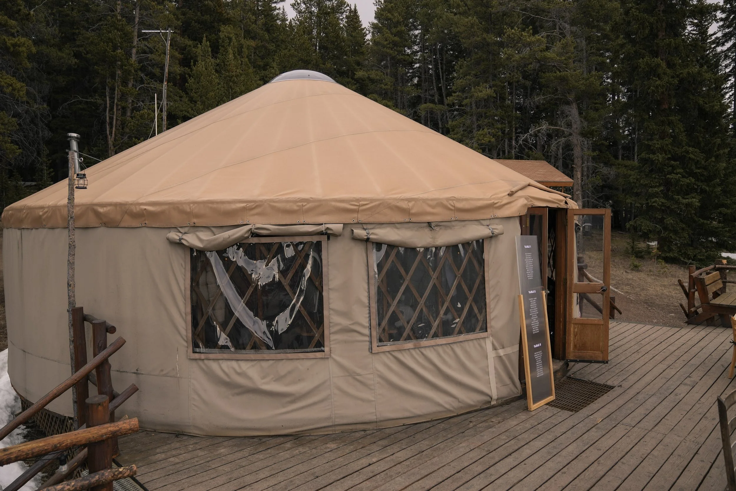A large beige circular tent with clear plastic window panels, set outdoors with a forest of evergreen trees in the background, wooden deck flooring, and a small blackboard sign outside.
