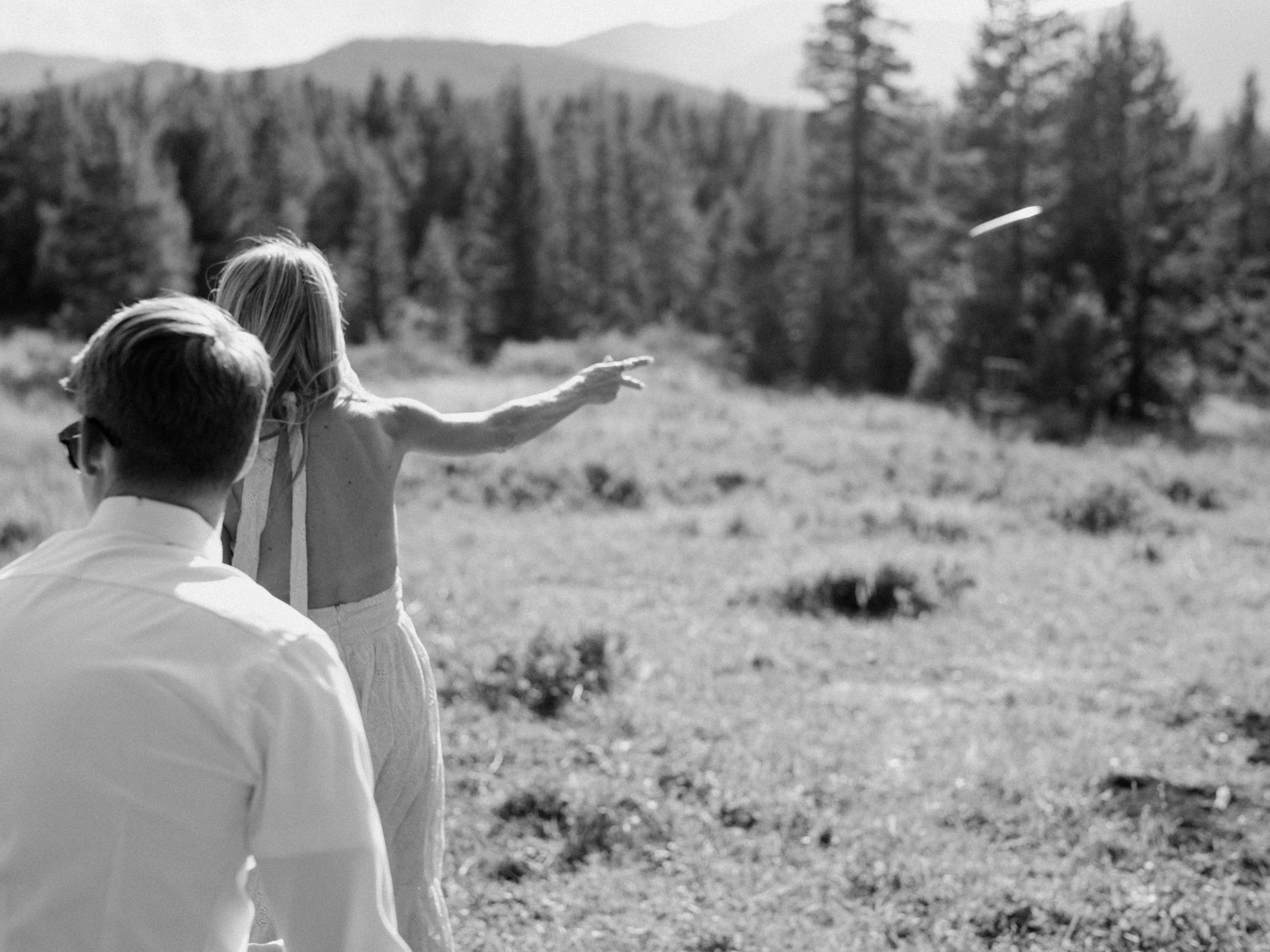 A black and white photo of a woman and:  a man in a field with trees and mountains in the background. The woman is pointing at something in the sky.