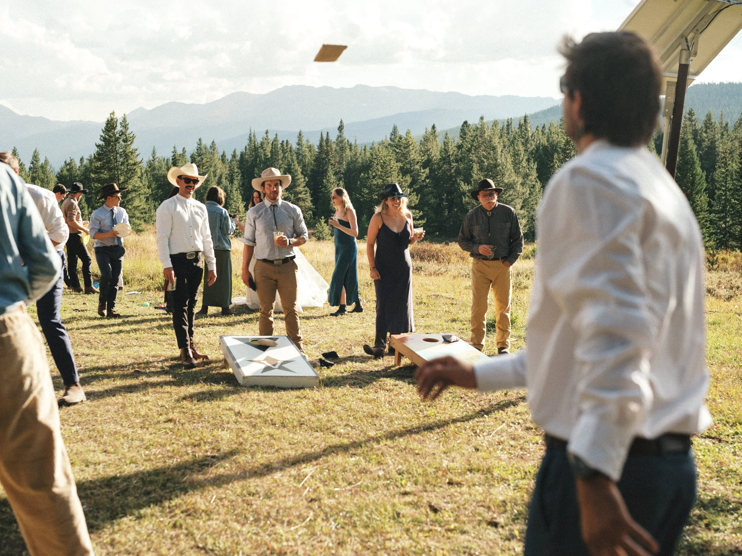 People participating in an outdoor cornhole game during a sunny day with mountains and trees in the background.