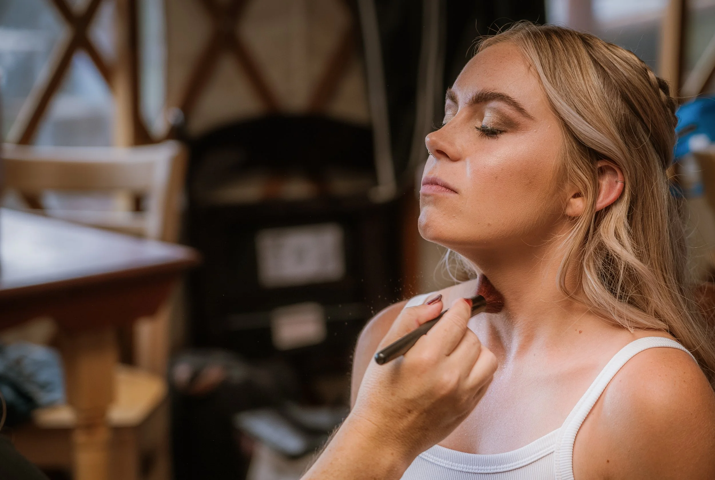 A woman with blonde hair and makeup, wearing a white tank top, has makeup applied by another person using a brush in a salon or makeup studio.