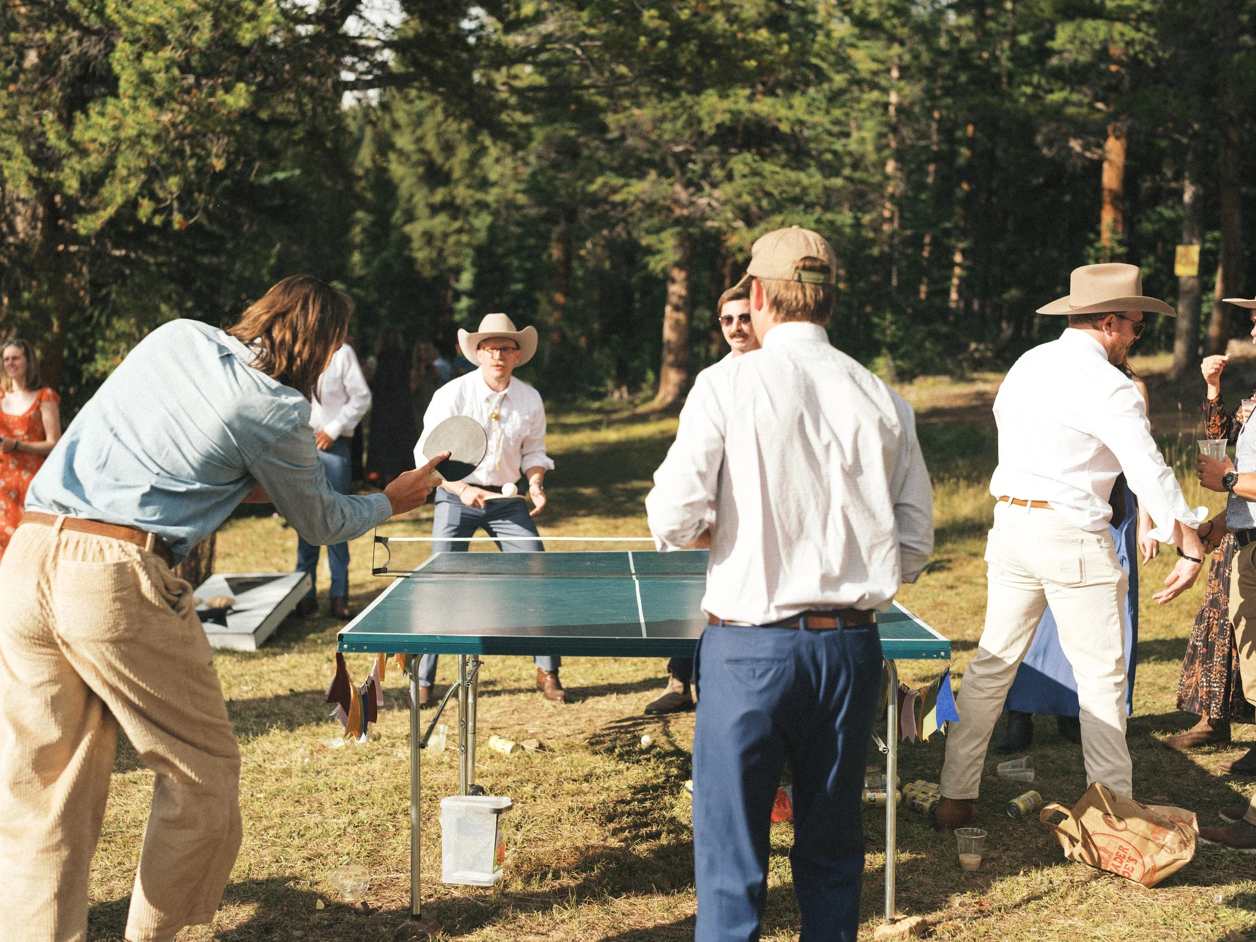 A group of people gathered outdoors in a wooded area playing ping pong, some dressed in Western-style clothing including hats and sunglasses, enjoying a casual social event.