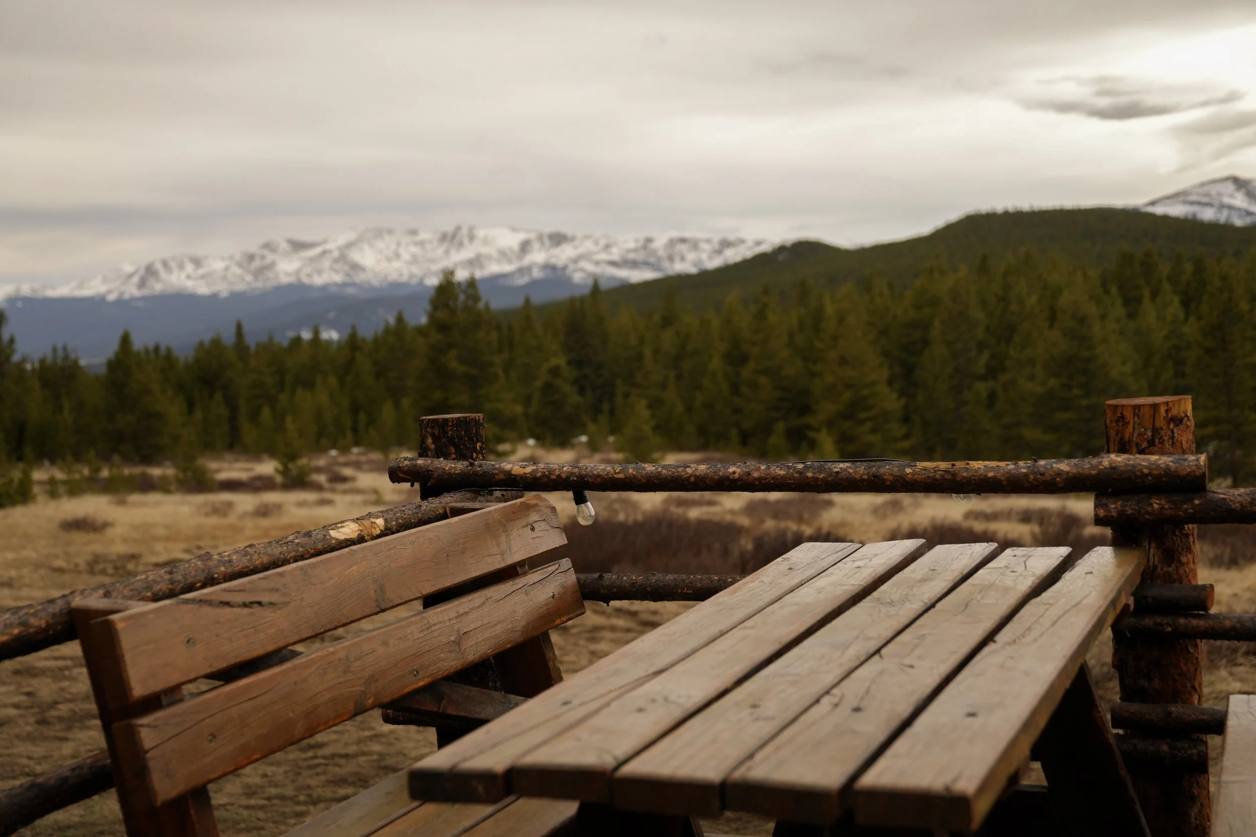 Wooden benches and railing in a scenic outdoor setting with a forest and snow-capped mountains in the background.