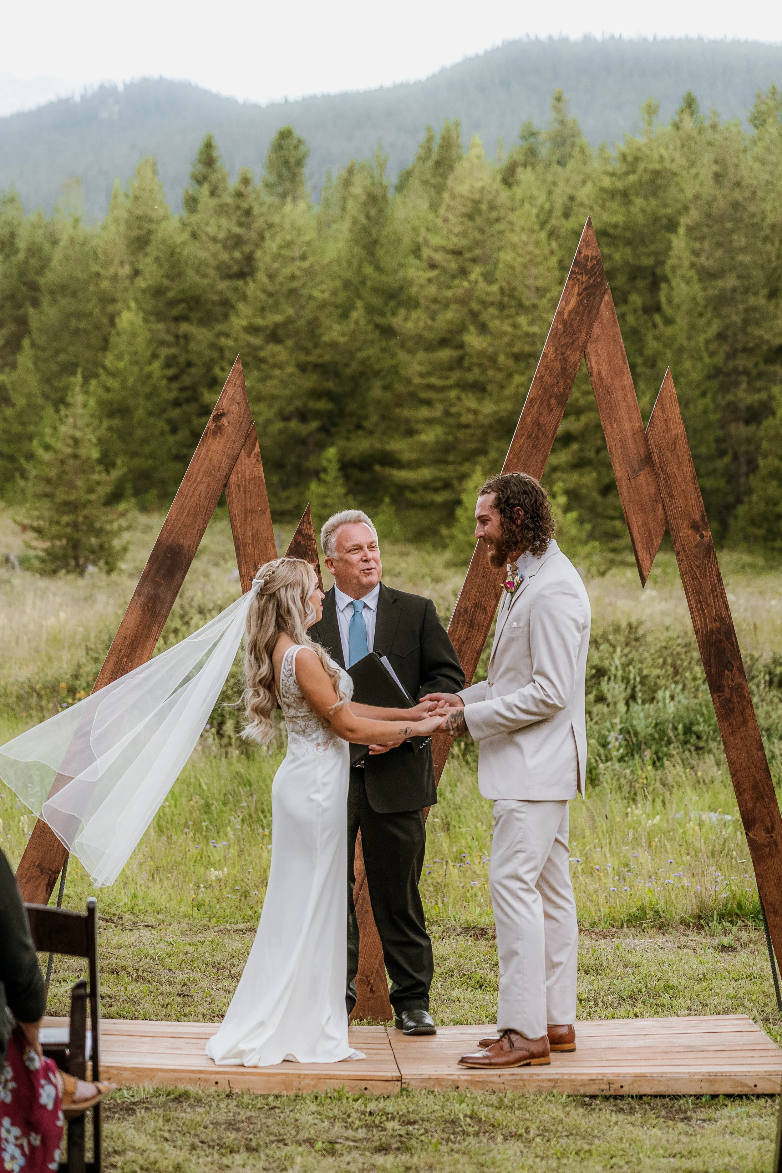 A couple getting married outdoors, holding hands and looking at each other, with an officiant and a mountain backdrop, under a large wooden wedding arch.