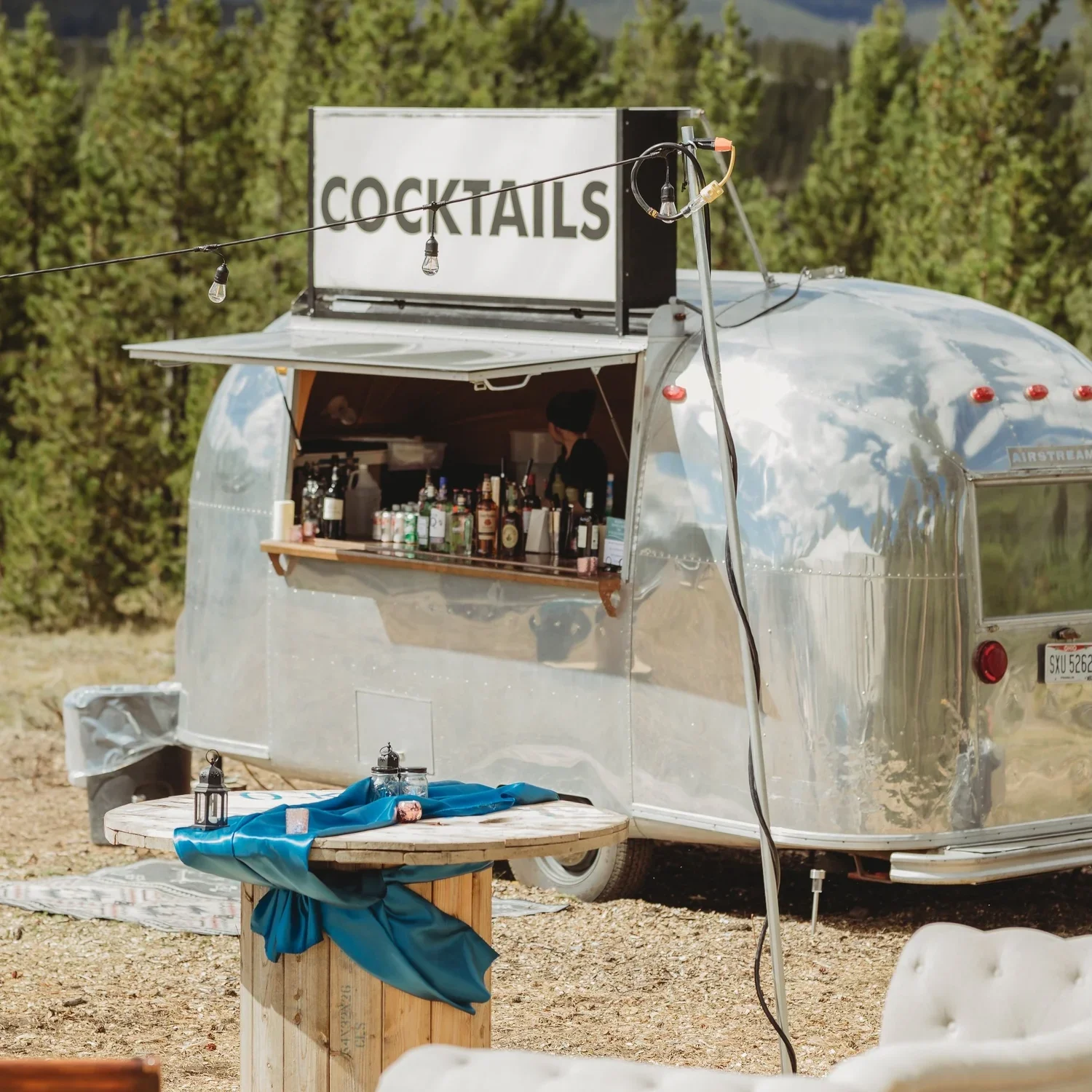A vintage silver Airstream trailer with a cocktail stand on top, displaying bottles of alcohol behind an open window, and a sign that says 'COCKTAILS' mounted above. There are string lights hanging above, and the setting is outdoors surrounded by trees.