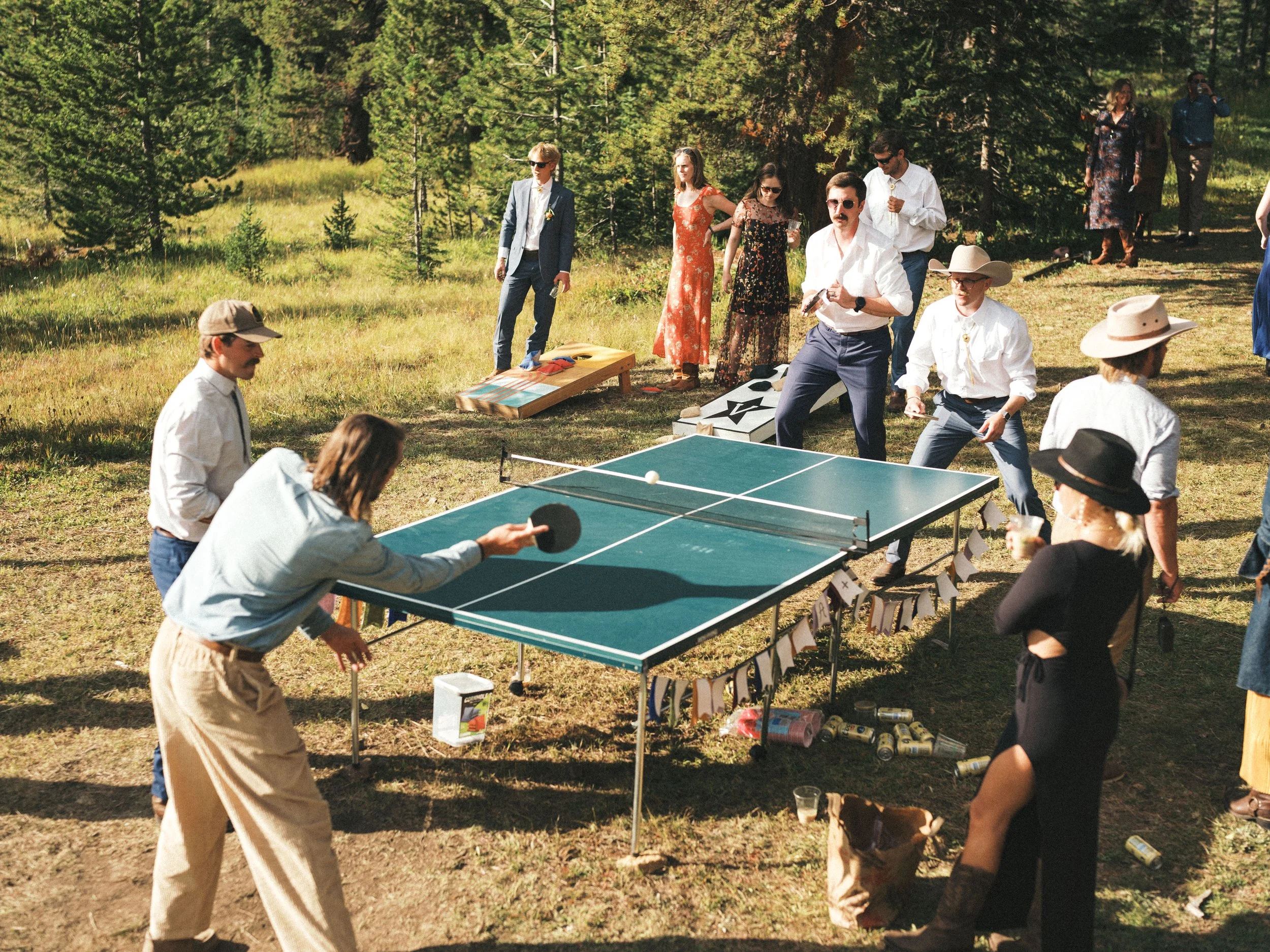 People playing ping pong outdoors in a grassy, wooded area, with some standing and watching and others actively engaged in the game.