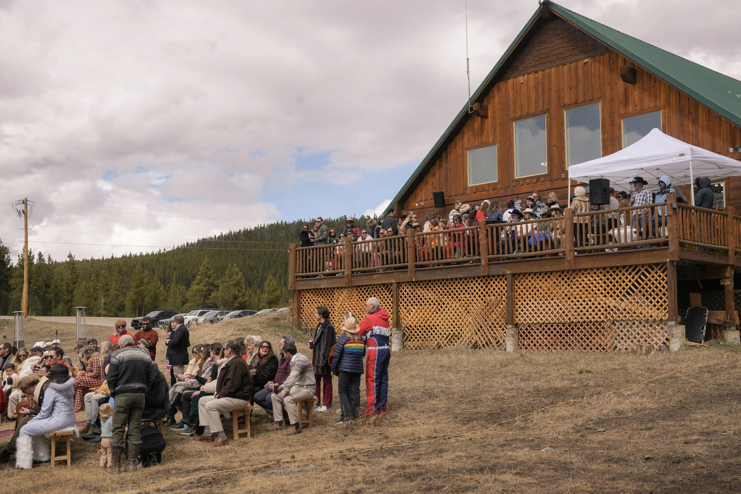 People gathered outdoors on a tiered wooden deck at a rustic lodge overlooking a wooded landscape under cloudy skies, with some chairs and a white canopy visible.