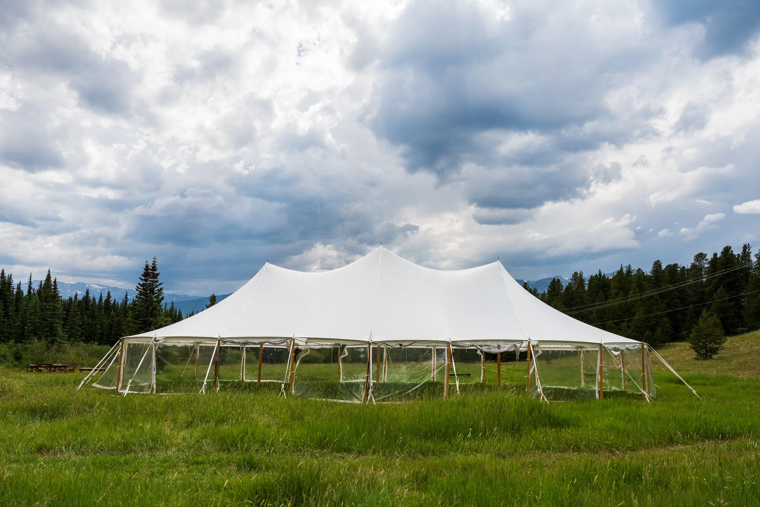 White event tent set up in a grassy field with forest and mountains in the background under a partly cloudy sky.