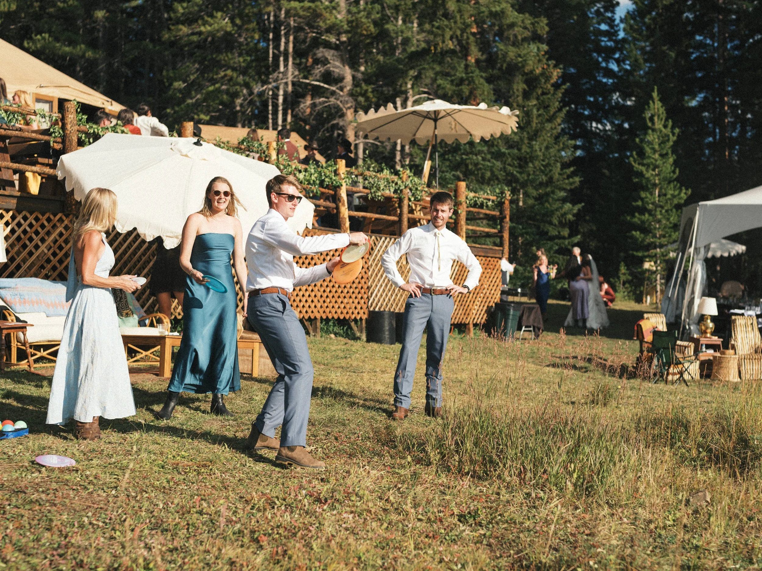 People at an outdoor event enjoying the sunny weather, some playing with a frisbee, surrounded by trees and a wooden terrace with umbrellas.
