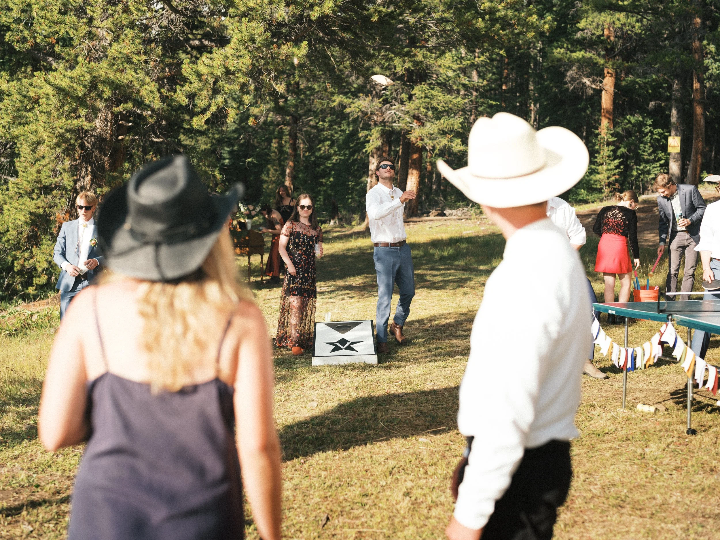 People playing cornhole outdoors in a forested area during daytime, with some watching and others participating in the game.
