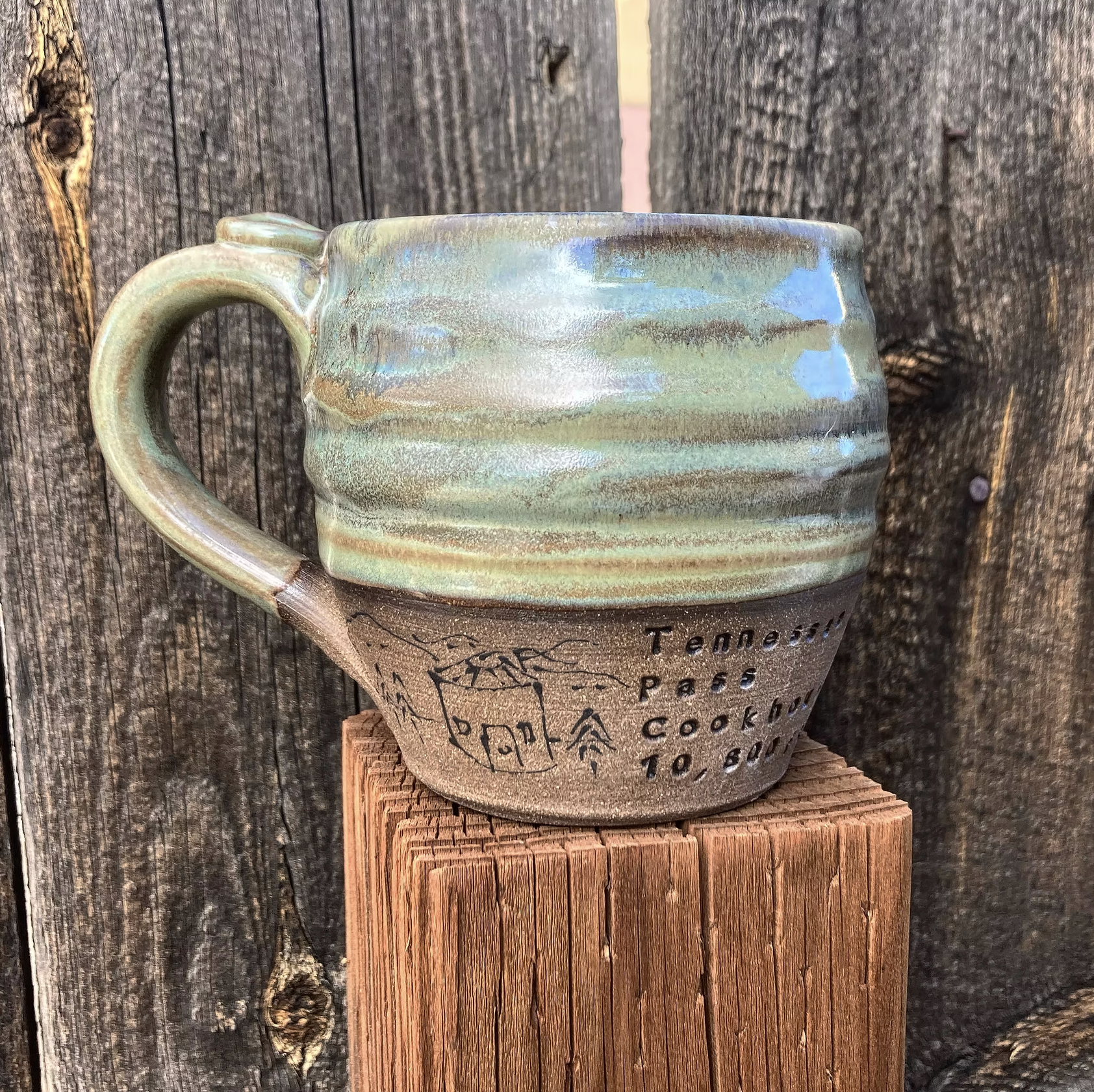 Close-up of a ceramic mug with a textured, multi-colored glaze, sitting on a wooden block against a dark, weathered wooden background.