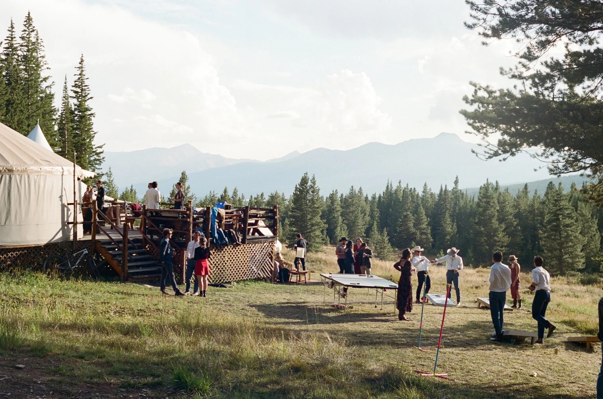 Outdoor gathering in a forested area with mountains in the background, people playing games, socializing, and a large white tent near a wooden platform.