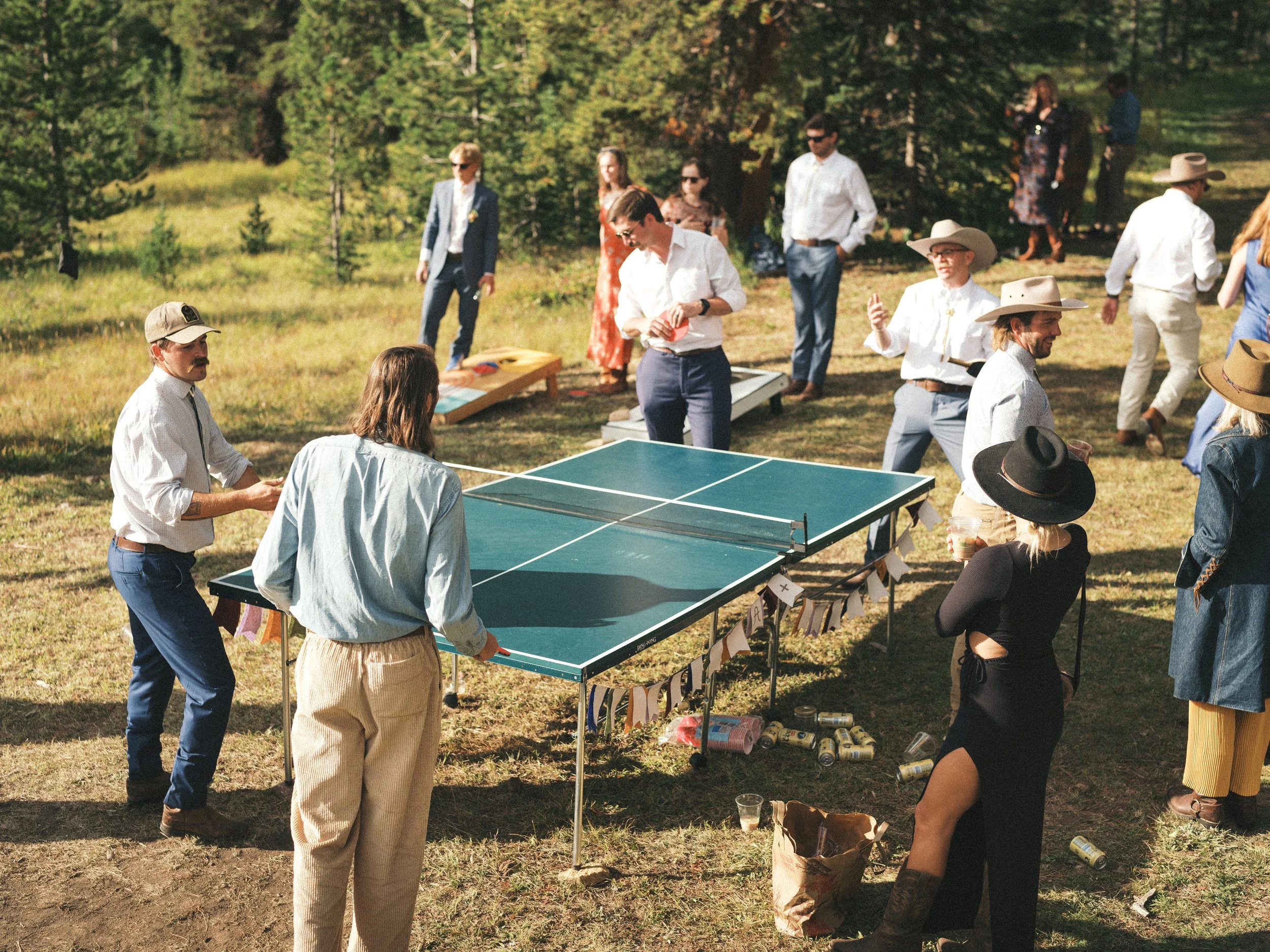 People gathered outdoors playing table tennis in a forested area during daytime, with some holding drinks and socializing.