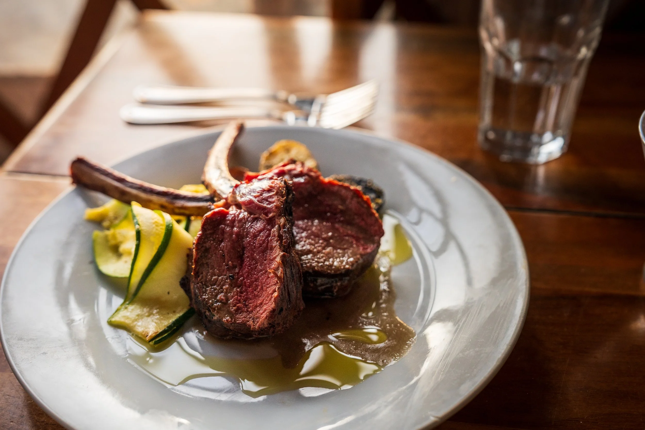 A plate with a cut of cooked red meat, zucchini noodles, and a yellow sauce, on a wooden table with a glass of water and silverware in the background.