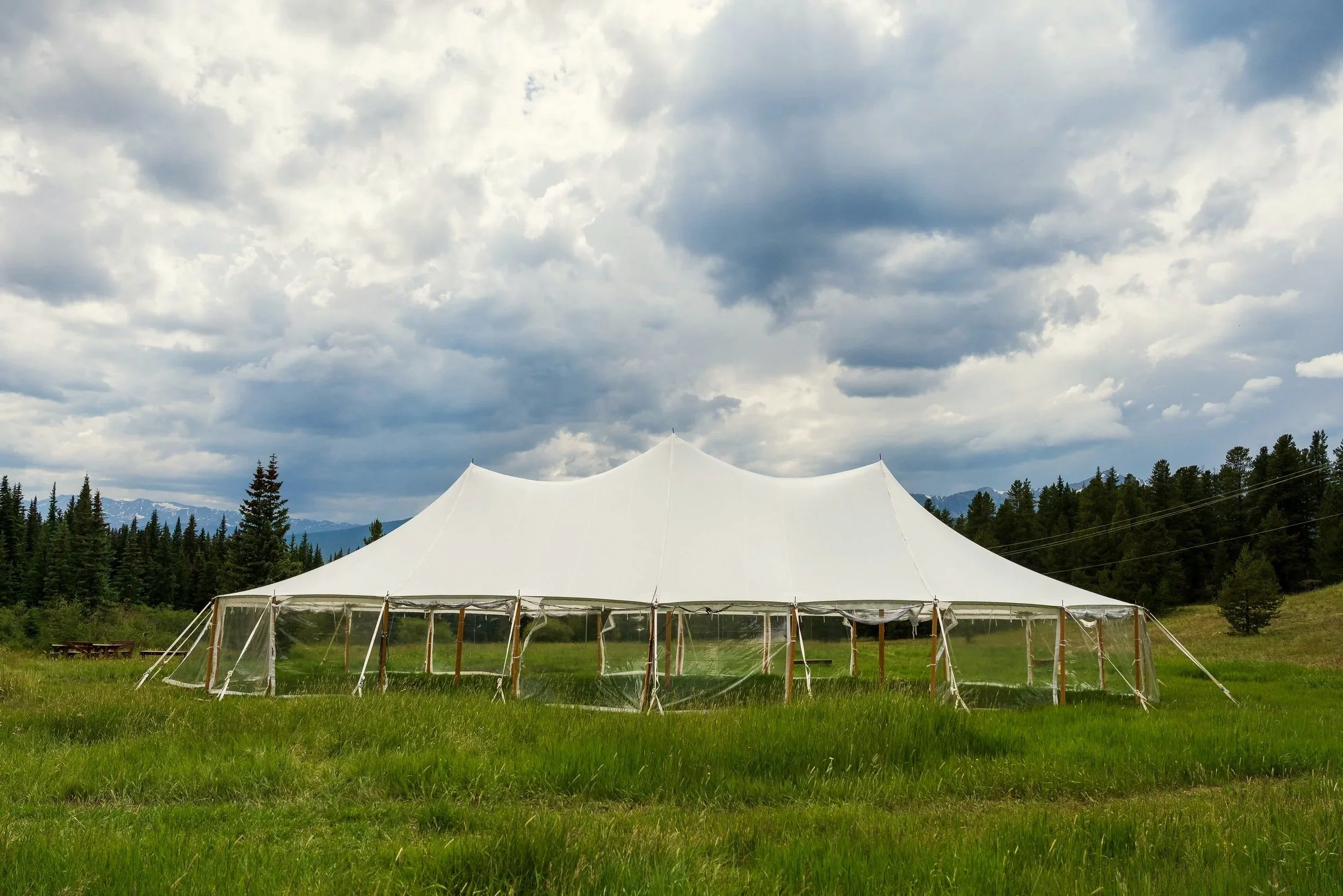 A large white tent set up in a green field with trees and mountains in the background under cloudy sky.