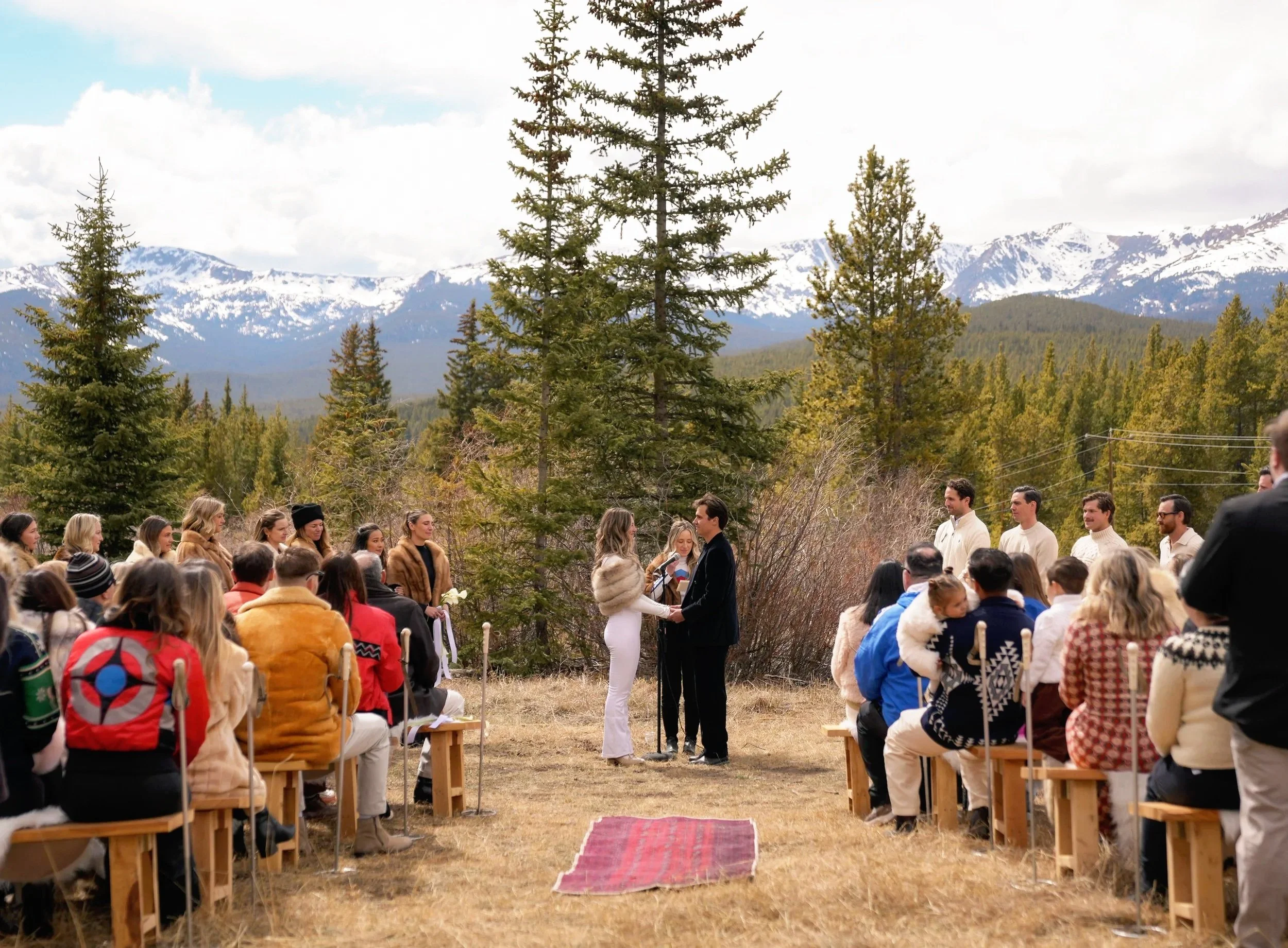 An outdoor wedding ceremony in a scenic mountainous area with coniferous trees and snow-capped peaks in the background. The ceremony is attended by numerous guests seated on wooden benches, facing a couple exchanging vows under an arch. The couple is holding hands and facing each other, with a woman officiating the ceremony.
