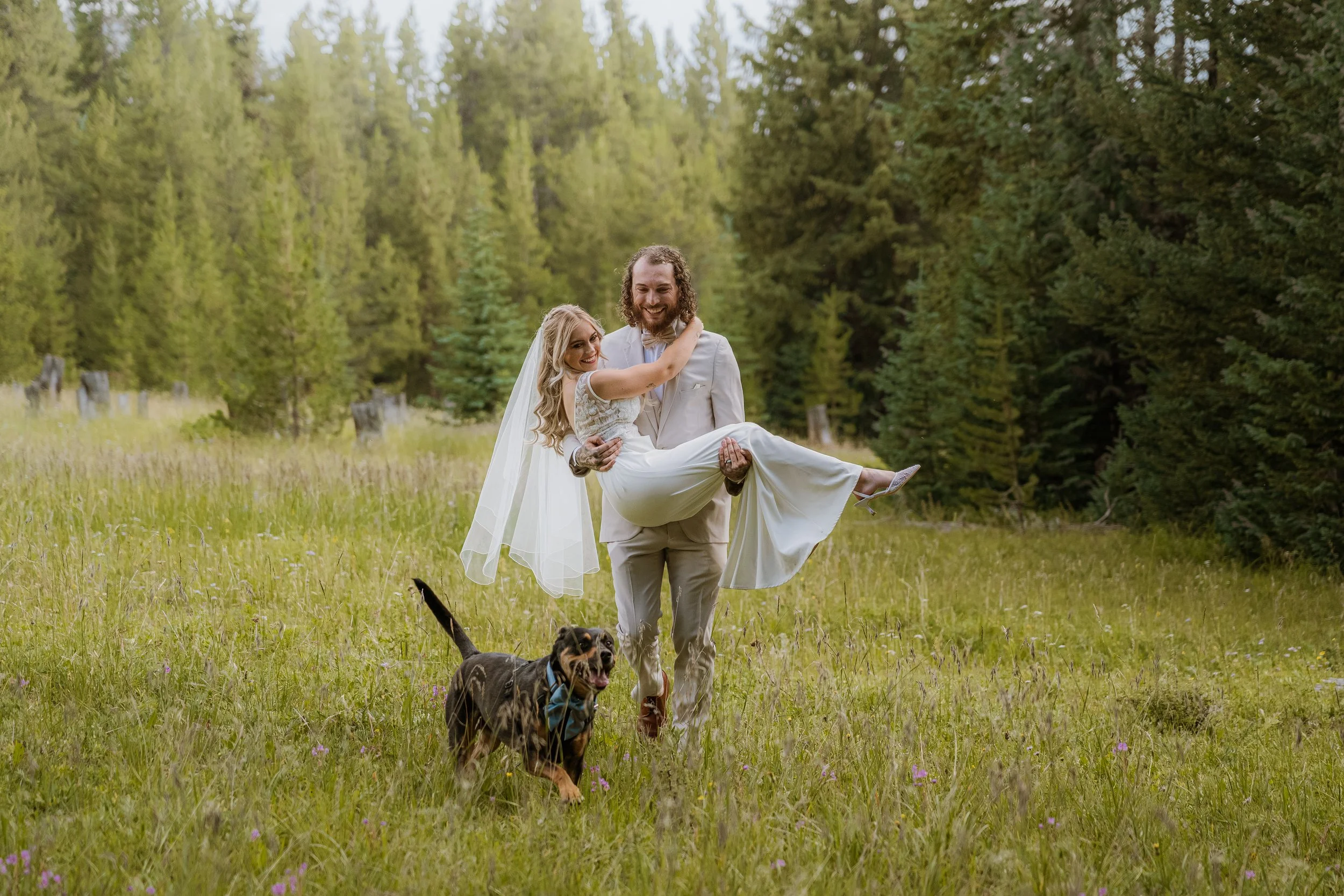 A bride and groom outdoors in a forest clearing, with the groom carrying the bride, both smiling. The bride is wearing a wedding dress and the groom a light-colored suit. Two dogs are running in the foreground.