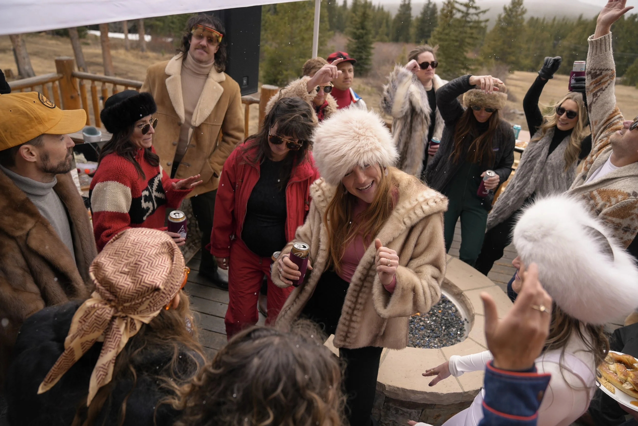 A group of people wearing winter clothes gathered outdoors, some holding drinks, enjoying socializing around a fire pit on a deck with trees in the background.