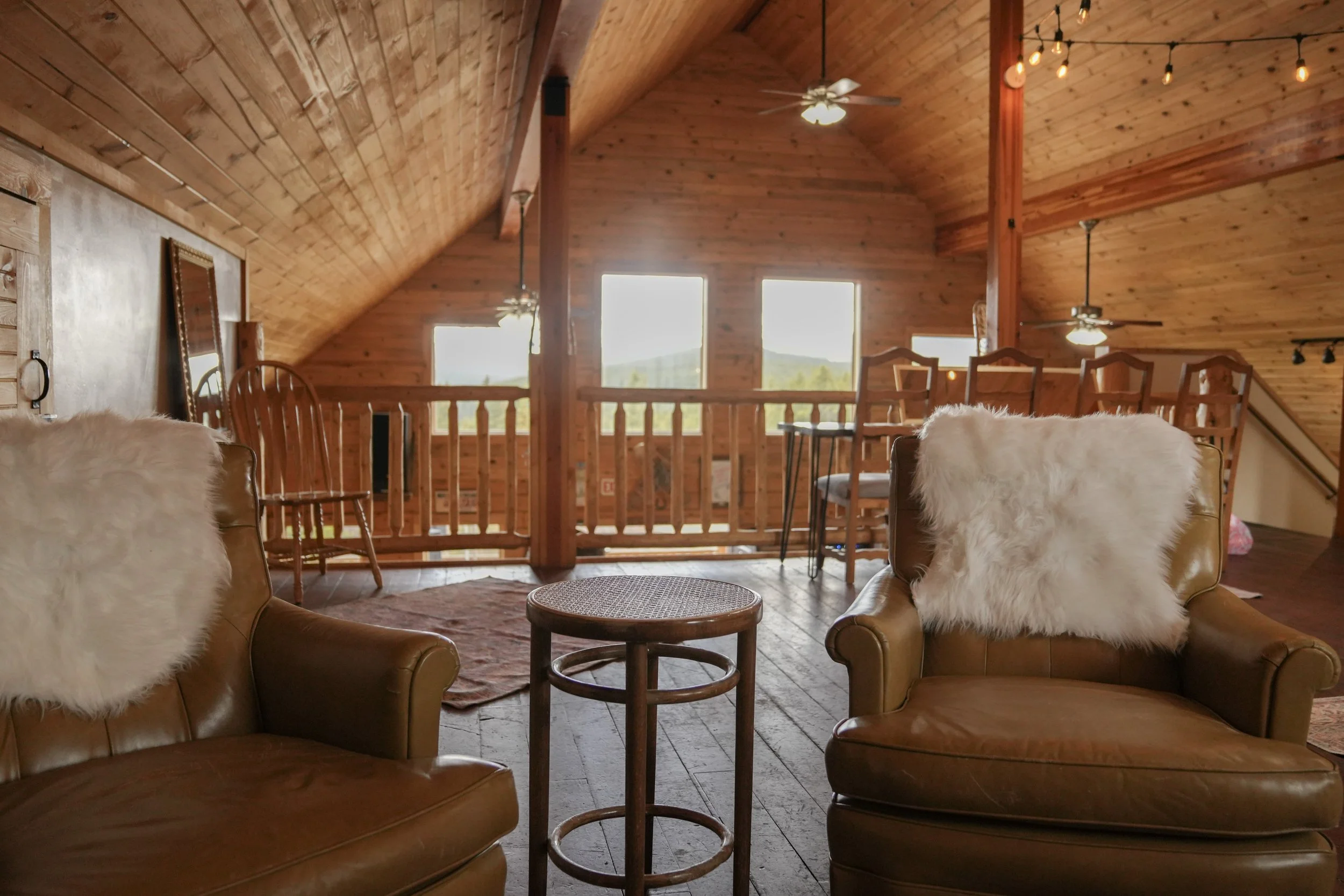 Interior of a cozy wooden cabin with leather armchairs, fluffy white pillows, a small round coffee table, and a railing overlooking the living space below. Large windows let in natural light, and string lights and ceiling fans decorate the ceiling.
