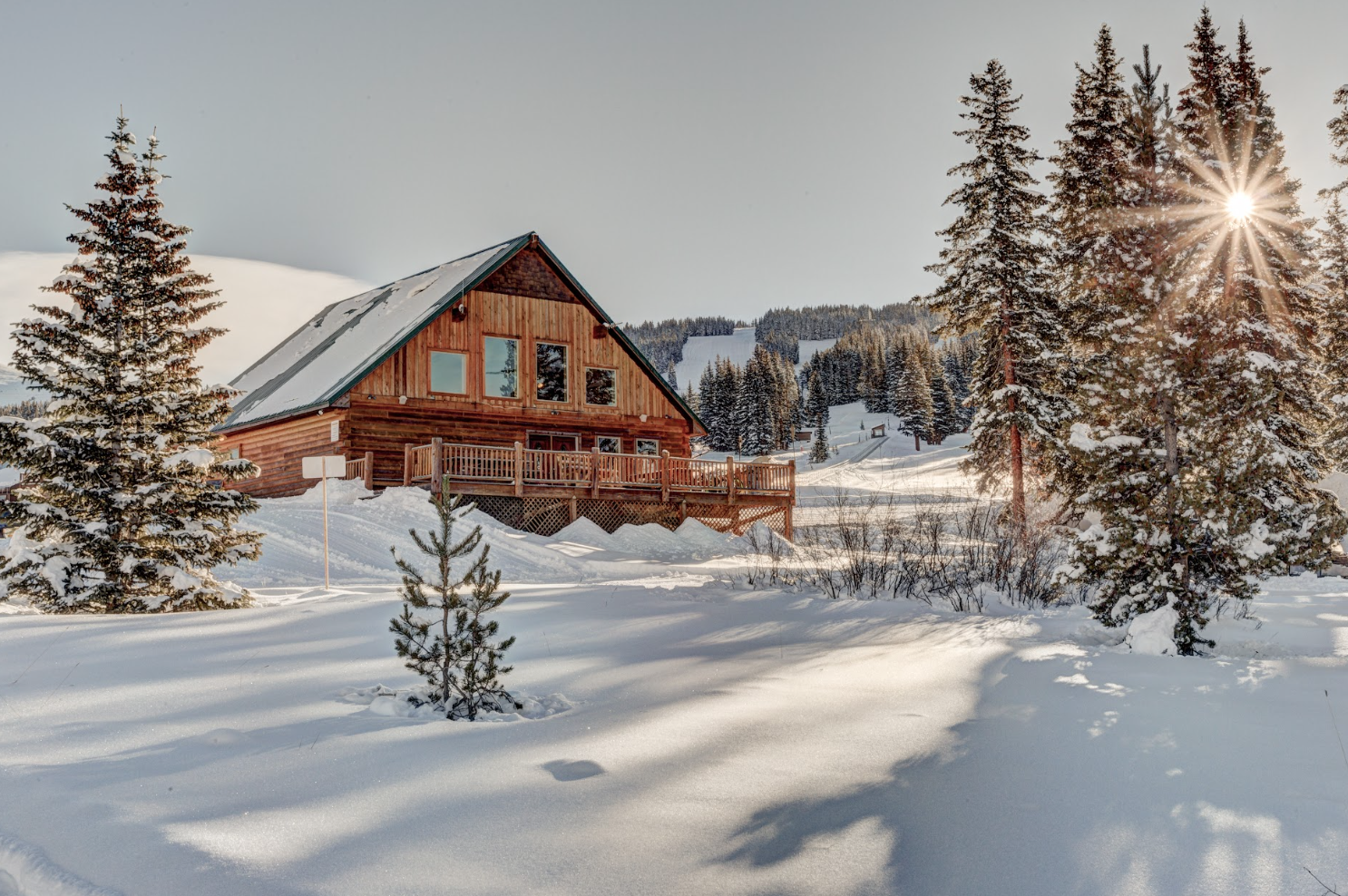 A wooden cabin in a snowy landscape with tall snow-covered evergreen trees and a bright sun shining through the trees.