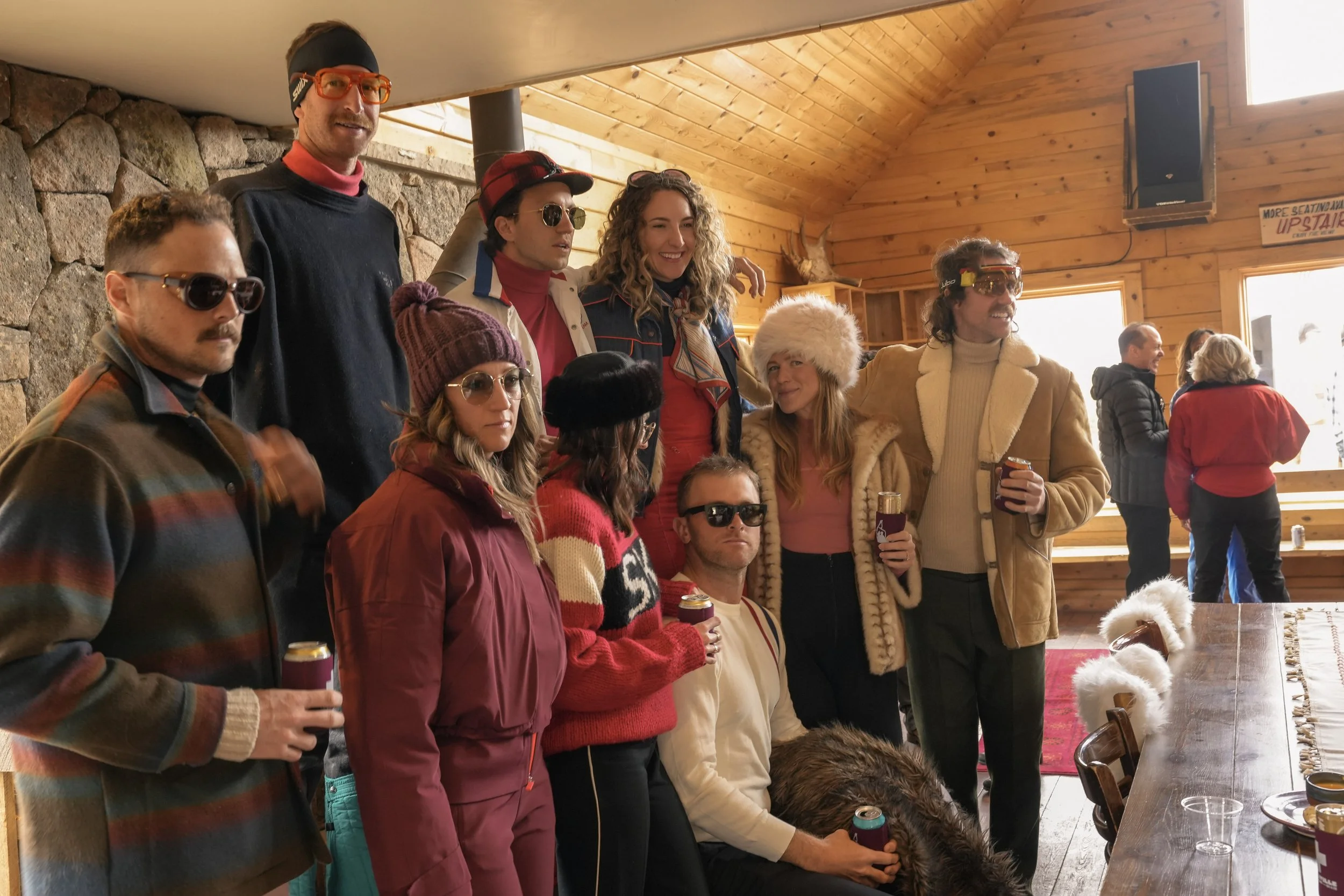 Group of people indoors at a gathering, some holding drinks, in a wood-paneled room with a stone wall, windows, and decorations including a mounted antler. Everyone appears to be dressed warmly, some wearing glasses and hats.