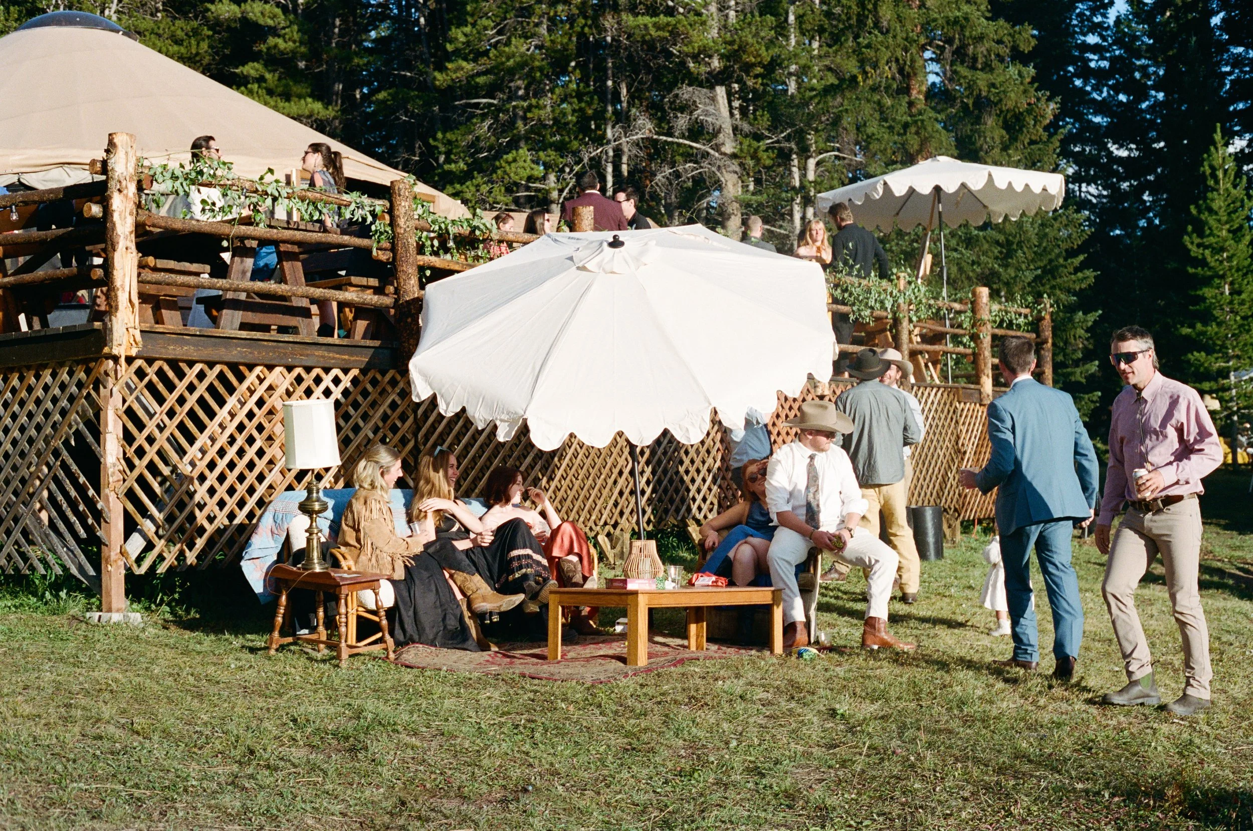 People gathered outdoors at a party or celebration, with some sitting on chairs and others standing, surrounded by trees with a wooden deck and umbrellas for shade.