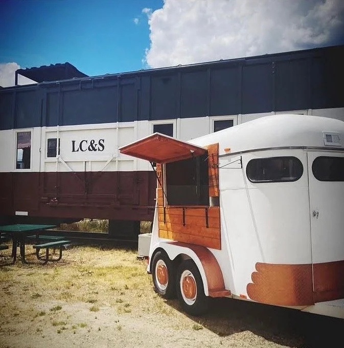 A white food truck with wood accents and a large window, parked next to a passenger train car with 'LC&S' markings, on a gravel lot with a picnic table and partly cloudy sky.