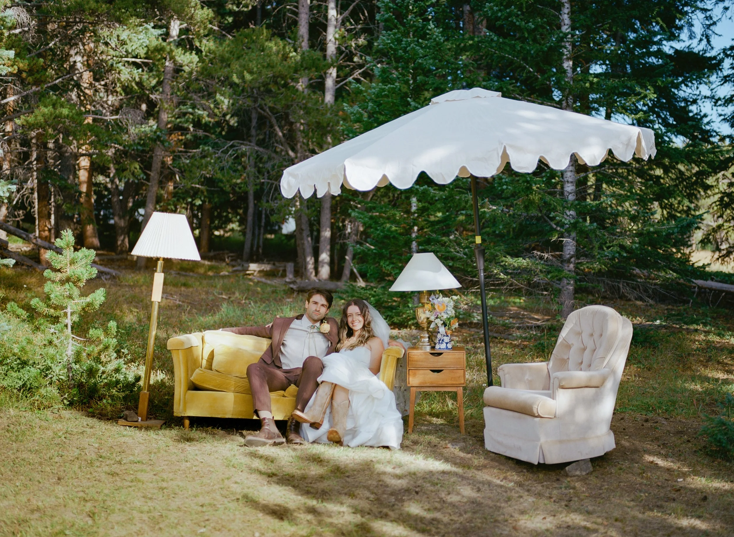 A couple sitting on a yellow sofa outdoors under a white parasol surrounded by greenery and outdoor furniture.