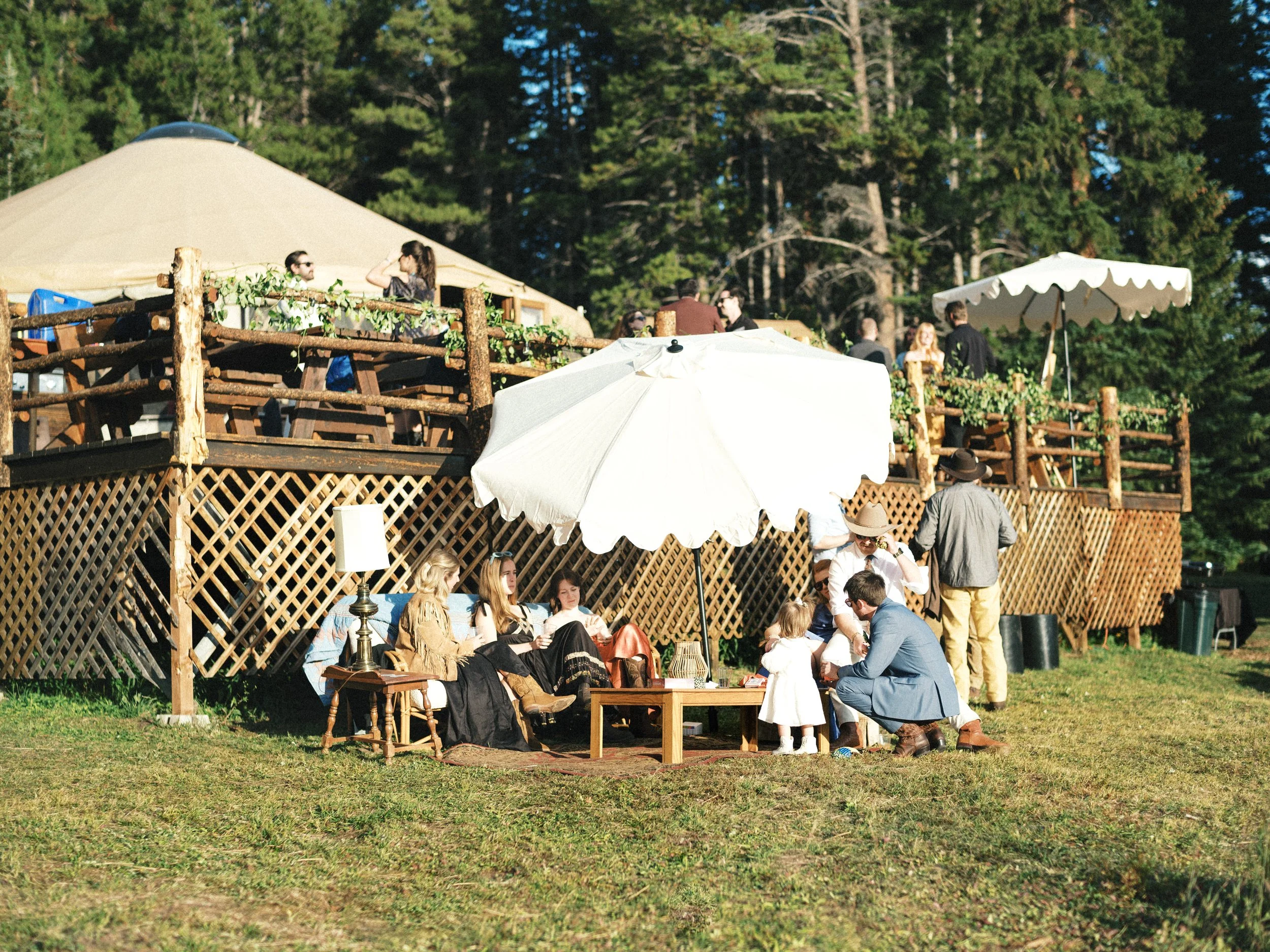 People gathering outdoors at a rustic wooden deck with a large beige tent, surrounded by trees, some sitting under a white umbrella, others standing and chatting during daytime.