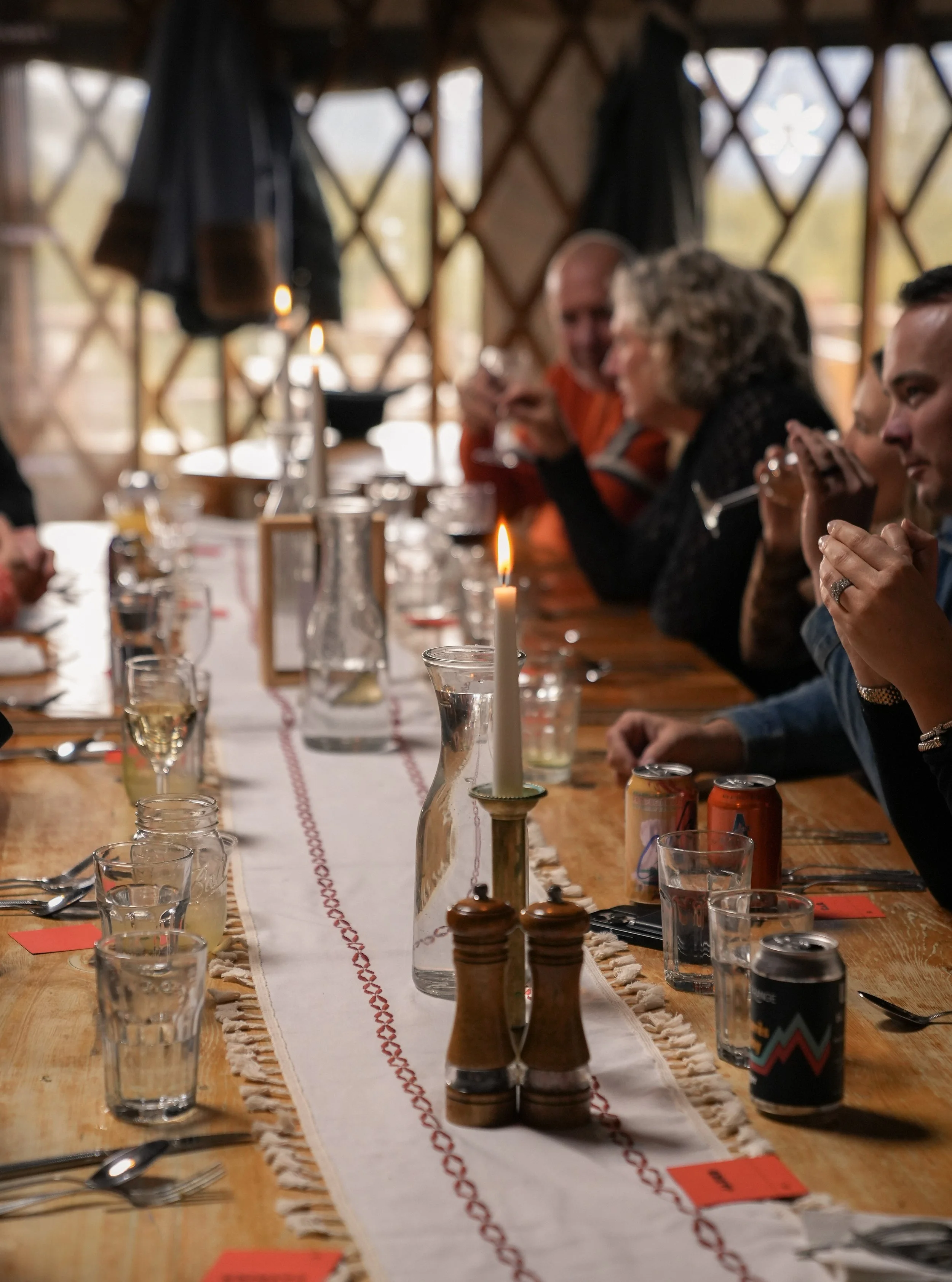 A long dining table set with glasses, plates, and cutlery, decorated with a white table runner with red embroidery, candles, and condiments, inside a wooden structure with lattice windows. Several people are seated along the table.