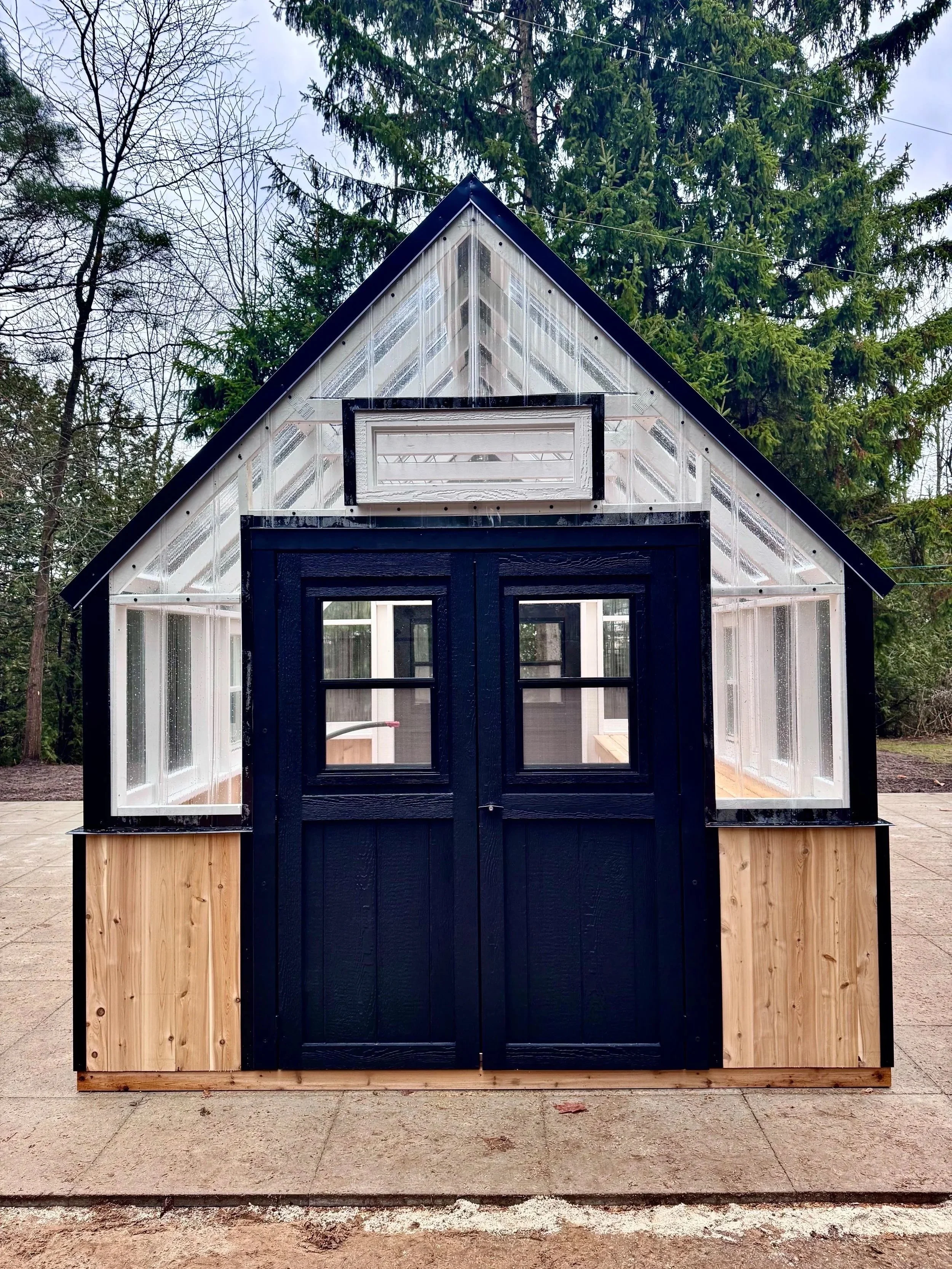 A small greenhouse with black double doors, wooden accents at the base, and large glass windows on the sides and roof, situated outdoors with trees in the background.