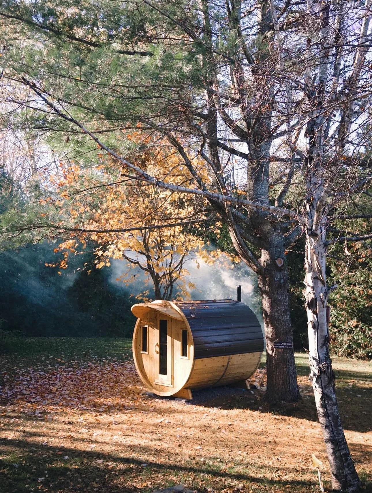 A small wooden barrel-shaped cabin with a black roof is situated among trees in a forested area during autumn.