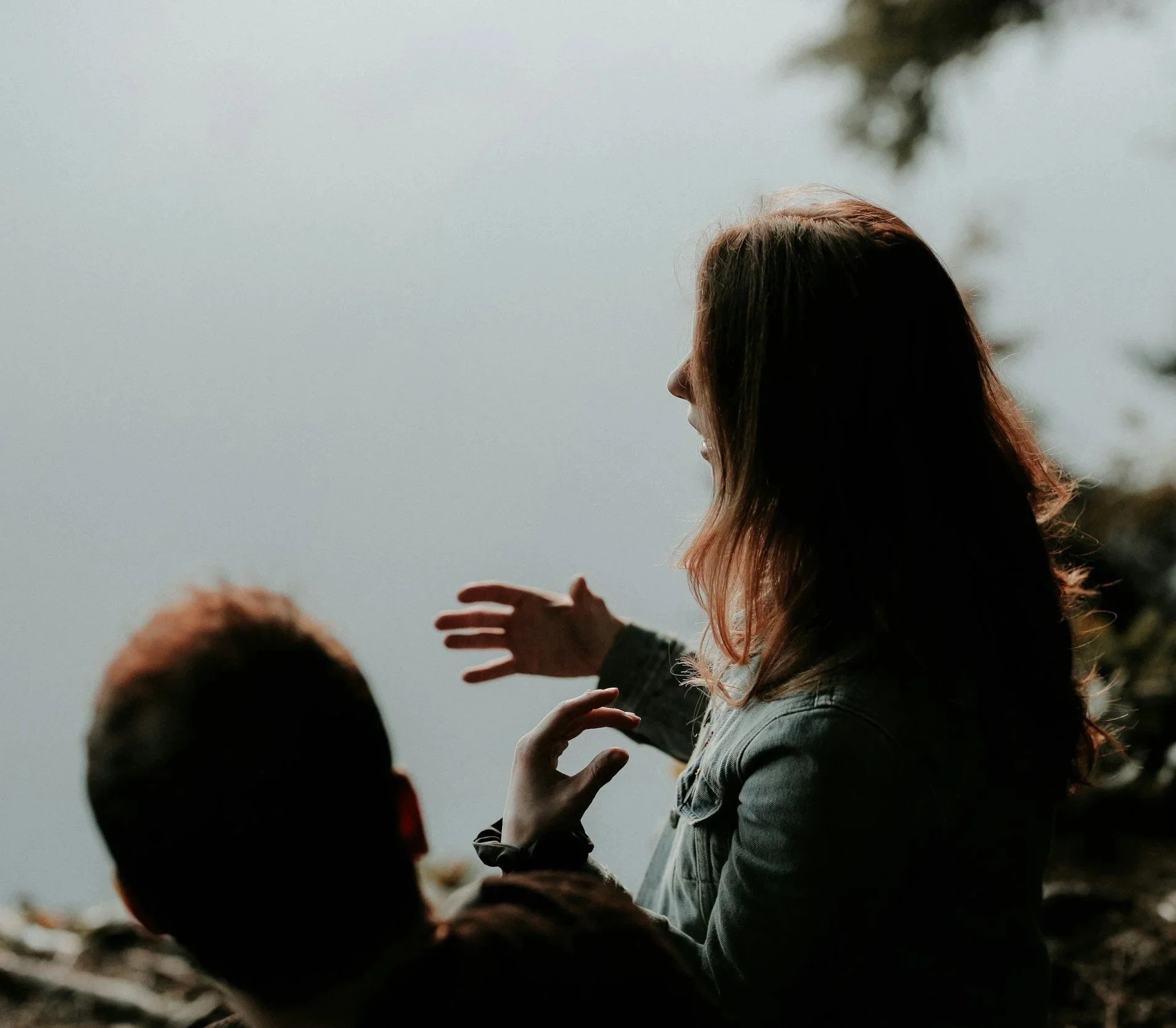 A woman with long hair standing near a body of water, with her arm extended, talking to a person whose head is visible in the foreground.