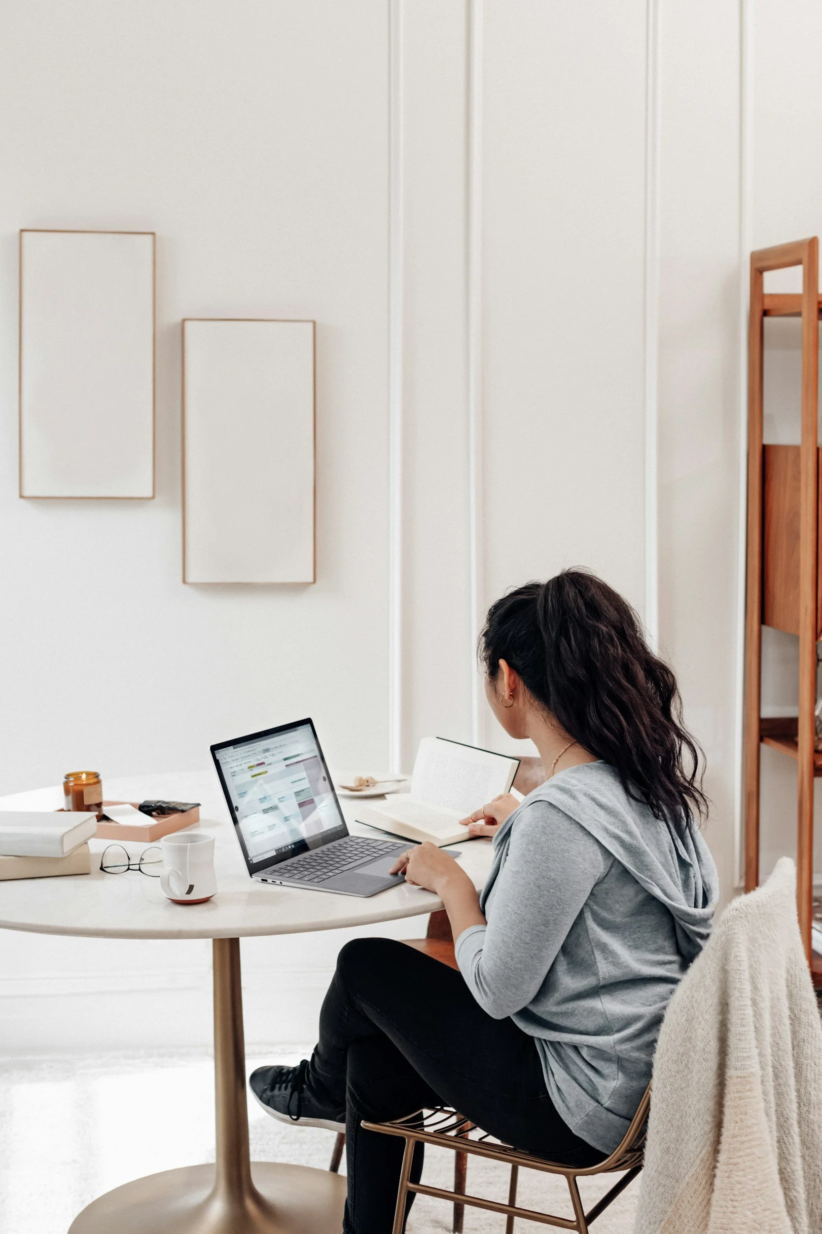 Woman sitting at a table, working on a laptop, with books and a mug on the table, in a bright, modern room with minimal decor.