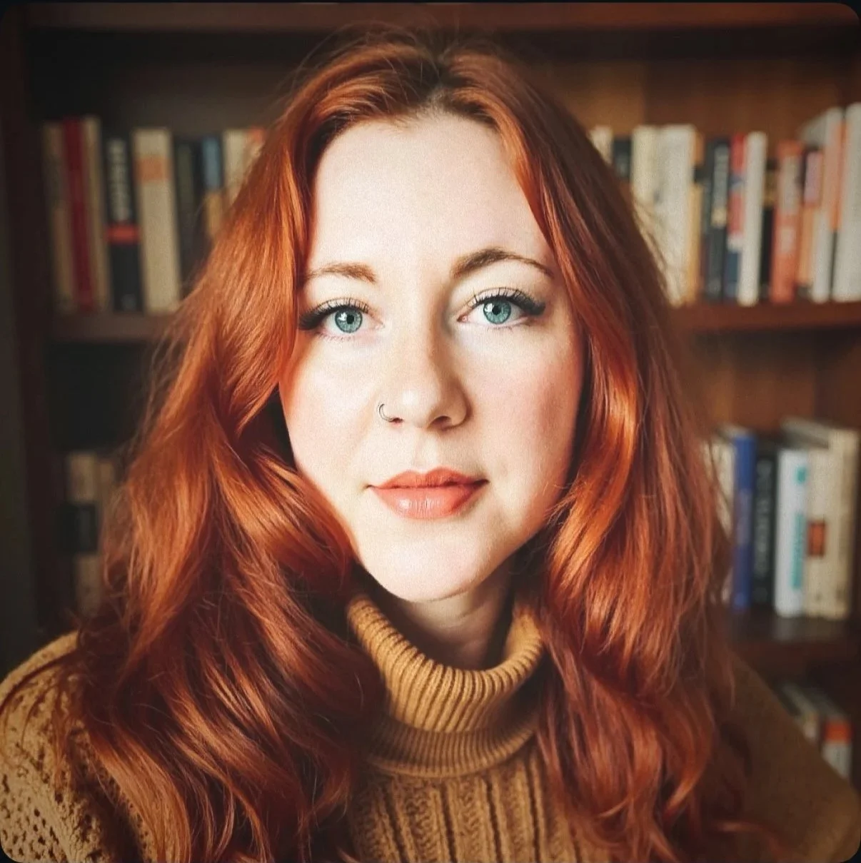 A woman with long, wavy red hair and blue eyes, wearing a mustard-colored turtleneck sweater inside a library.