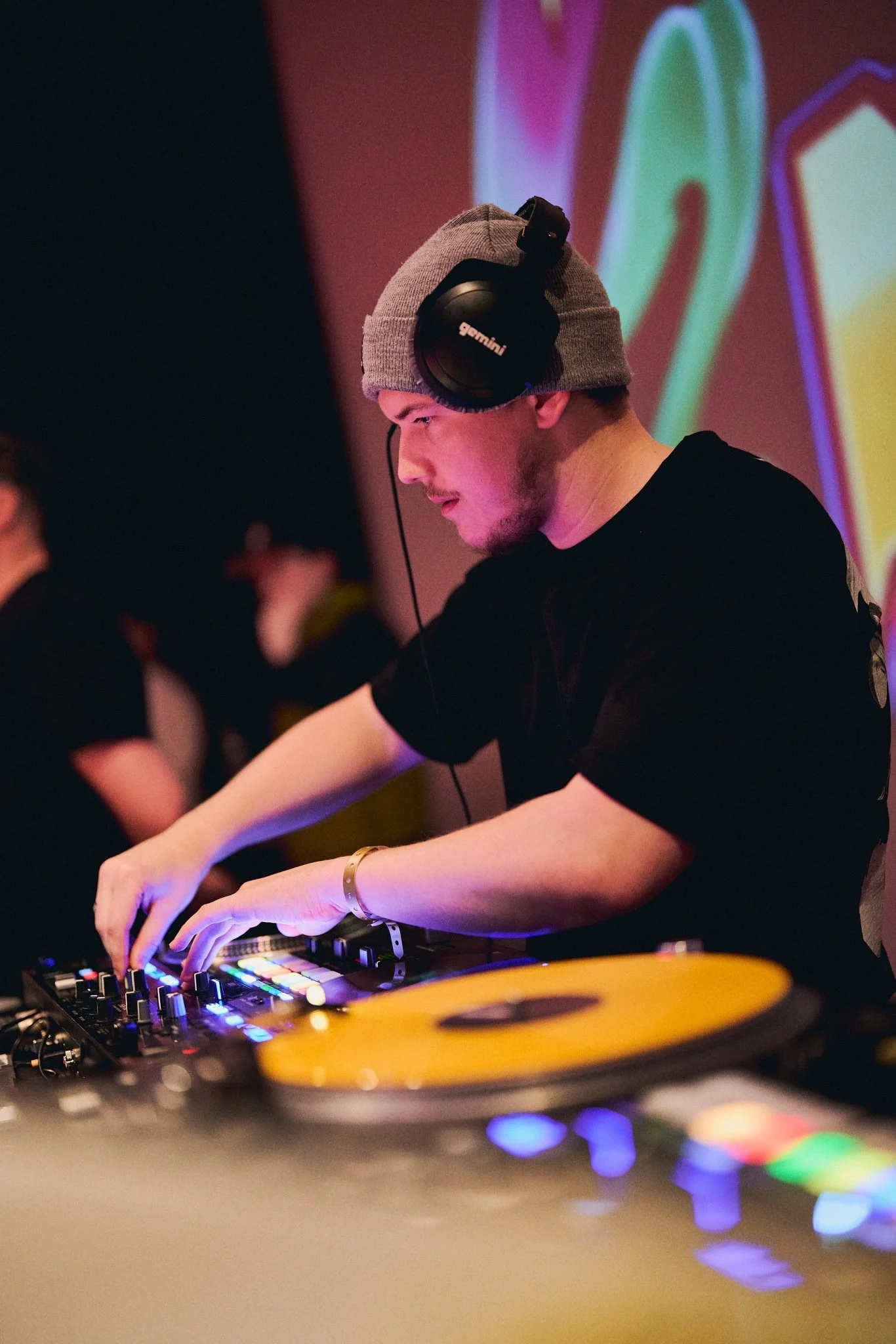 A young man DJing at a club with colorful visuals in the background, wearing headphones, a gray beanie, and a black shirt.