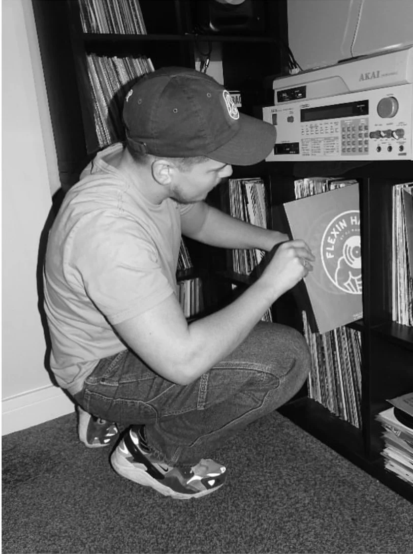 A person crouches down to look at and handle a record album on a shelf, wearing a baseball cap, t-shirt, jeans, and sneakers, surrounded by shelves filled with vinyl records and electronic equipment.