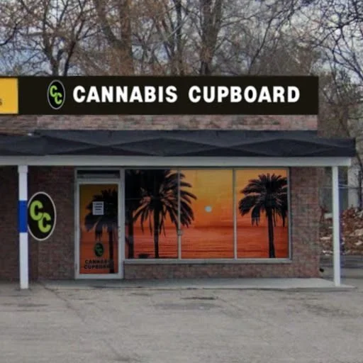 storefront with a sign reading 'CANNABIS CUPBOARD' and a large window depicting a sunset with palm trees.