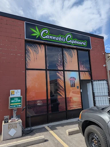 Storefront with a sign reading 'Cannabis Cupboard' accented by a marijuana leaf, large reflective windows showing palm trees and sunset, parking lot with a tire inflator station and a gray vehicle.