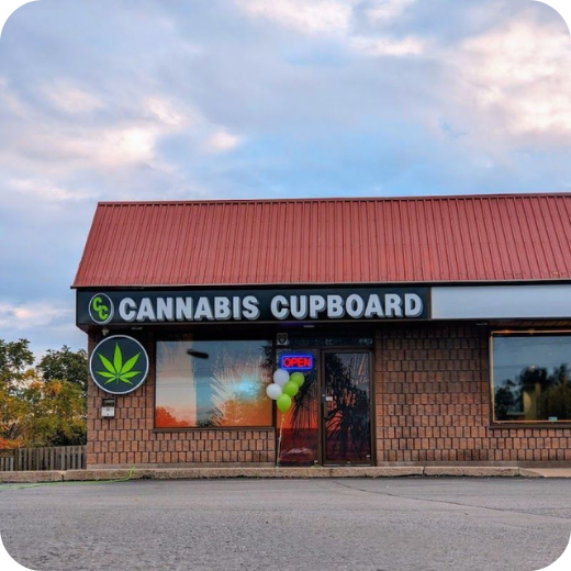 storefront with sign reading 'CANNABIS CUPBOARD', marijuana leaf logo, balloons on door, open sign, brick building with red roof, and cloudy sky