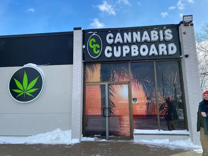 Exterior of Cannabis Cupboard store with signage featuring a green cannabis leaf, a sunset reflection on glass doors, and snow on the ground in an urban area.