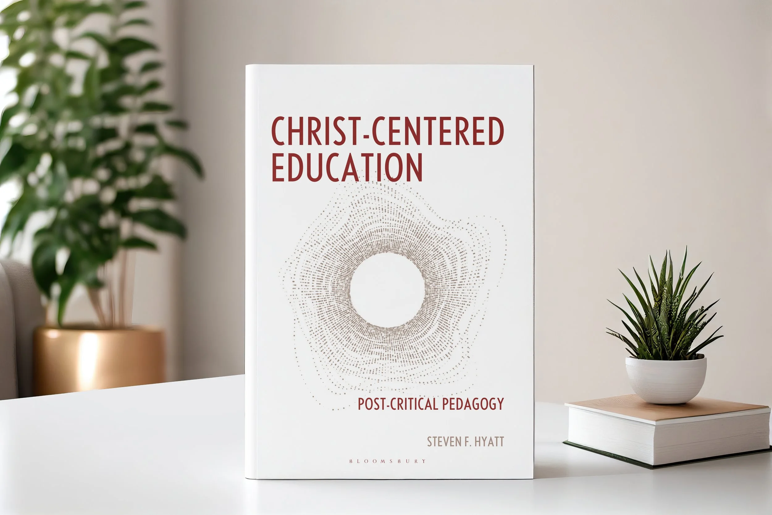 A book titled 'Christ-Centered Education: Post-Critical Pedagogy' by Steven F. Hyatt is standing upright on a white table. There are two potted plants, one with broad leaves in a gold pot to the left and a cactus in a white pot on top of another book to the right. The background is a plain, neutral-colored wall.
