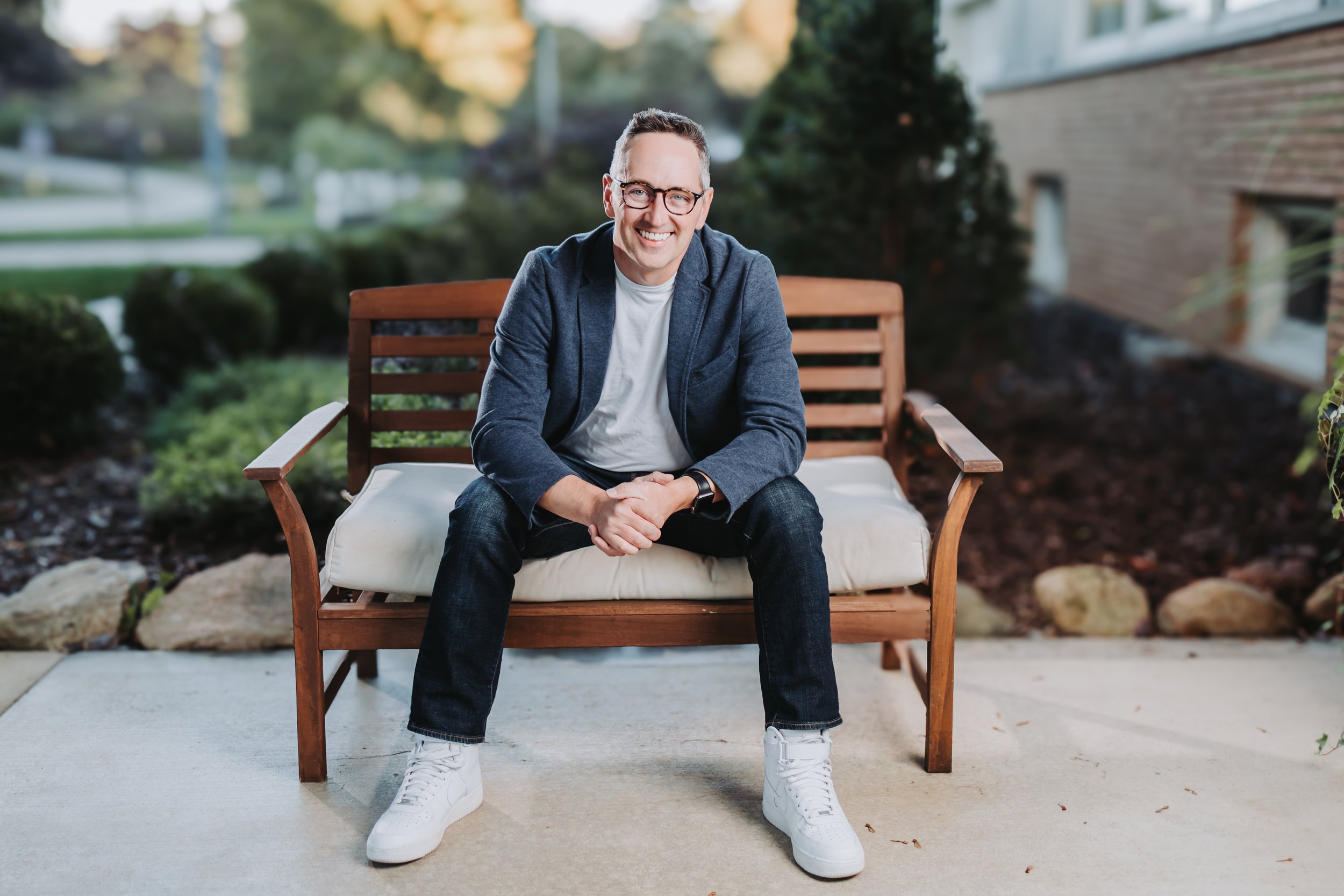 A man in glasses, a dark blazer, white t-shirt, jeans, and white sneakers sitting on a wooden outdoor bench, smiling at the camera, with a backyard garden and house in the background.