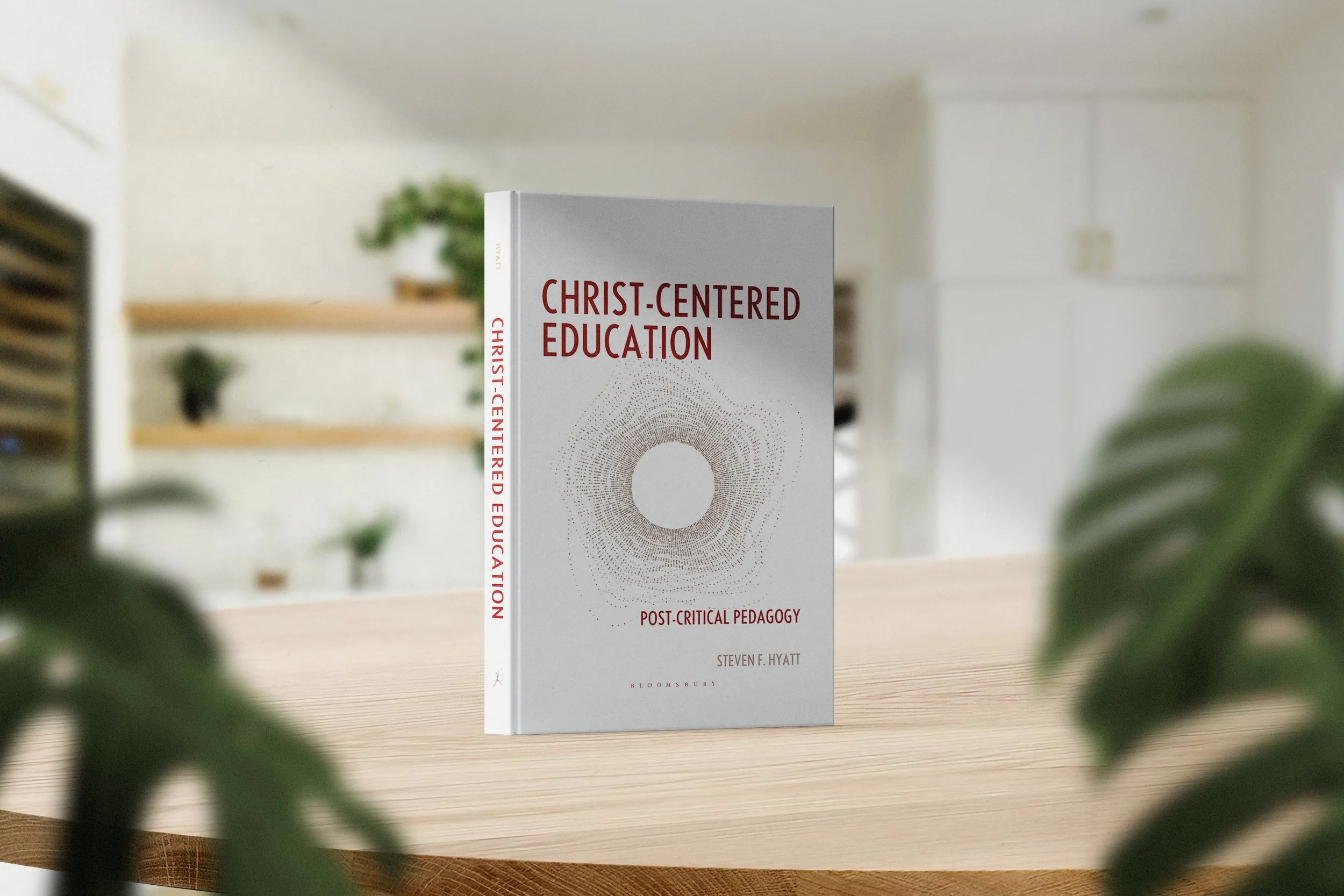 A book titled "Christ-Centered Education: Post-Critical Pedagogy" by Steven F. Hyatt, placed on a light wooden table with a blurred bookshelf and potted plants in the background.