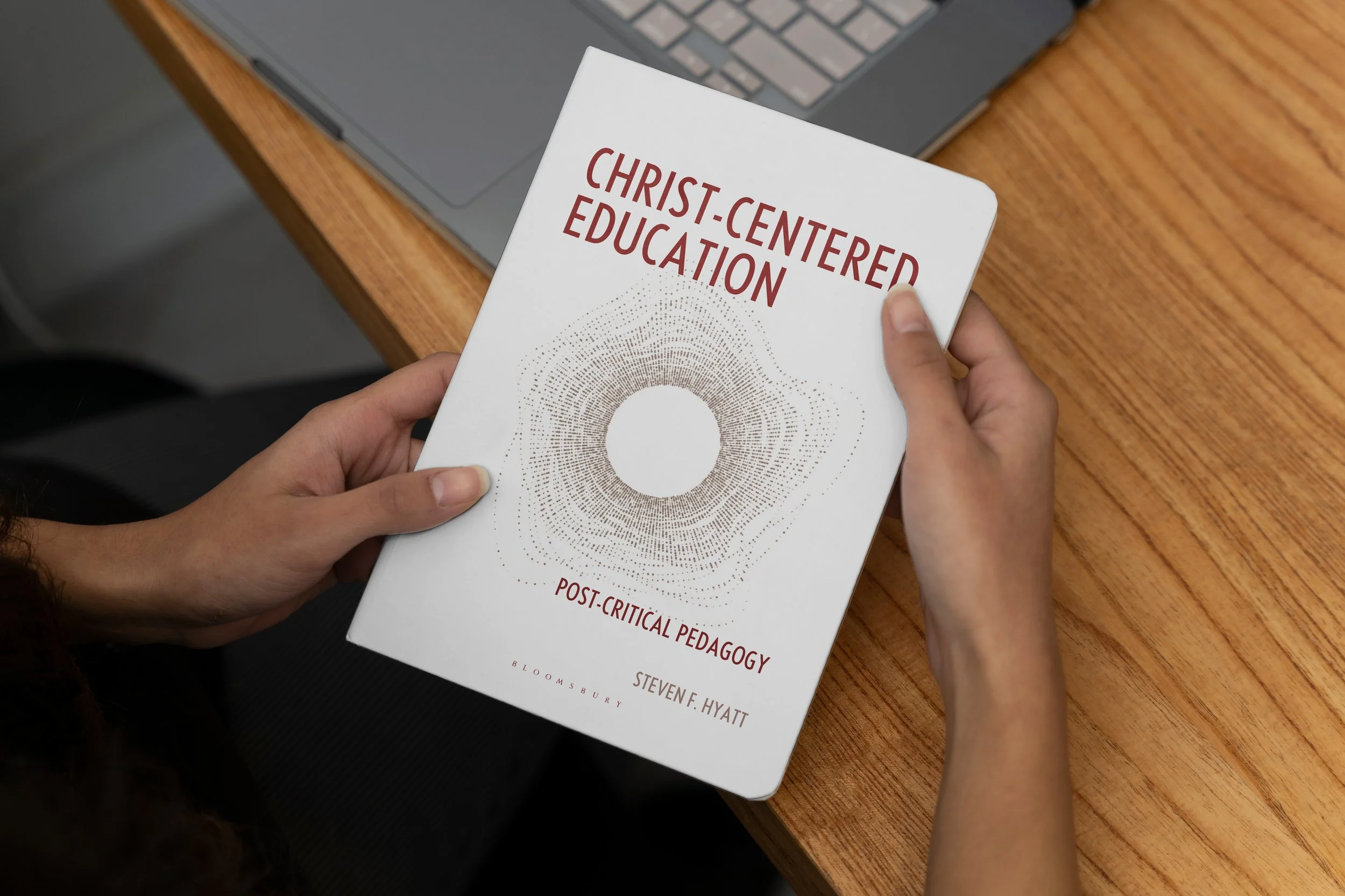 Person holding a book titled "Christ-Centered Education: Post-Critical Pedagogy" by Steven F. Hyatt, with a laptop on a wooden desk in the background.