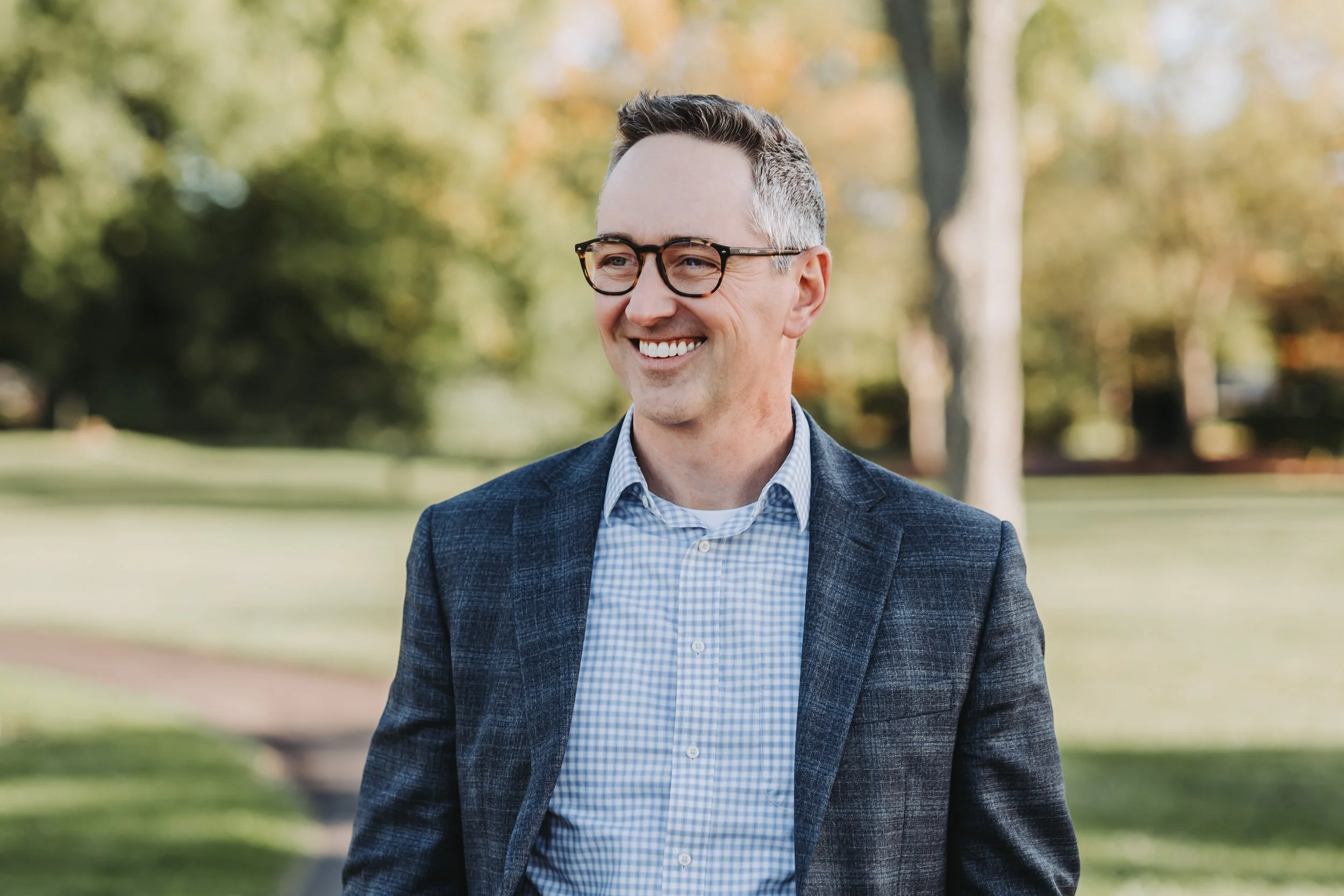 A smiling man wearing glasses, a checkered shirt, and a blazer outdoors in a park with trees and grass.